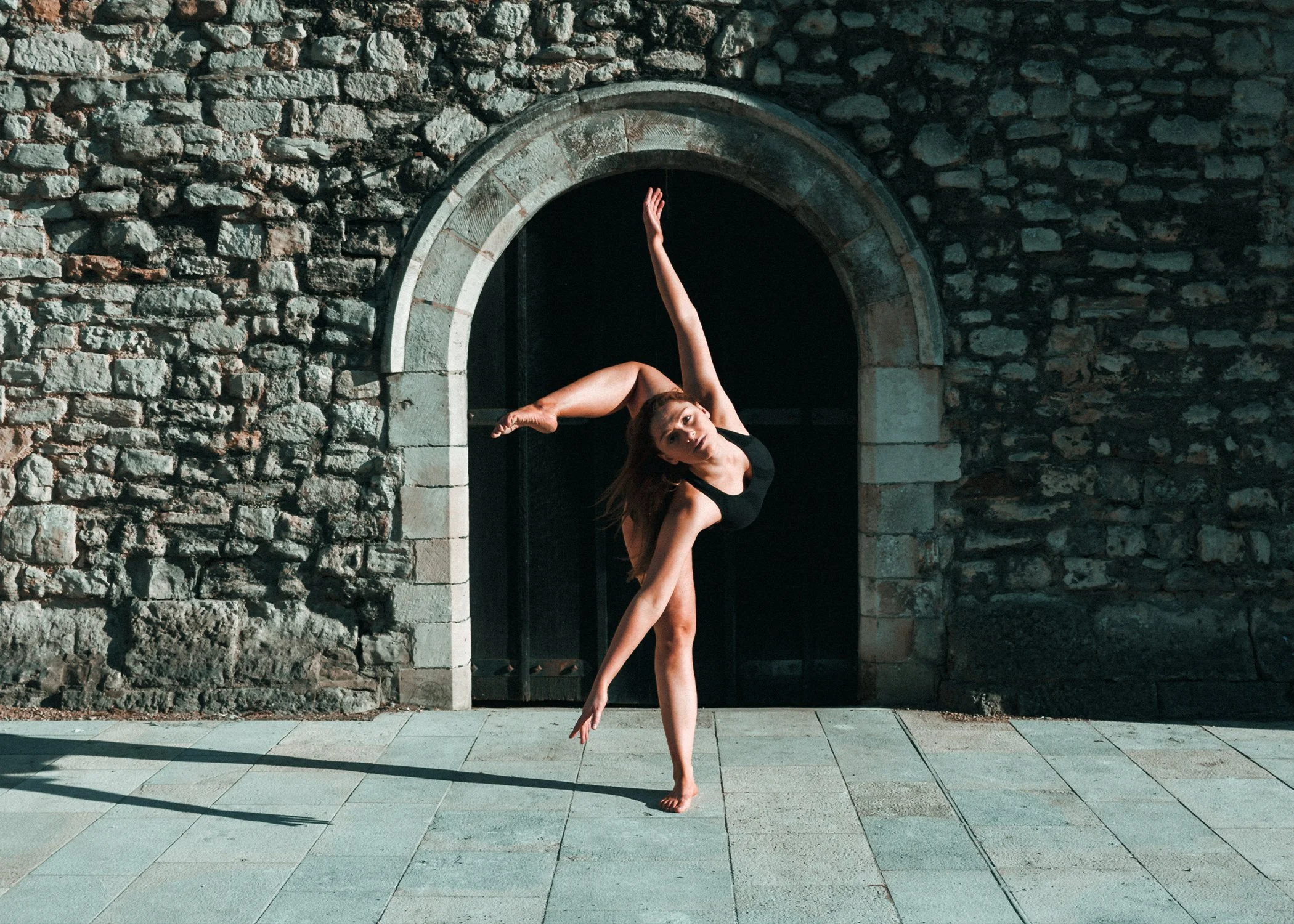 A woman performing a ballet pose outdoors against a stone wall with an arched doorway, standing on a paved surface with shadows cast on the ground.