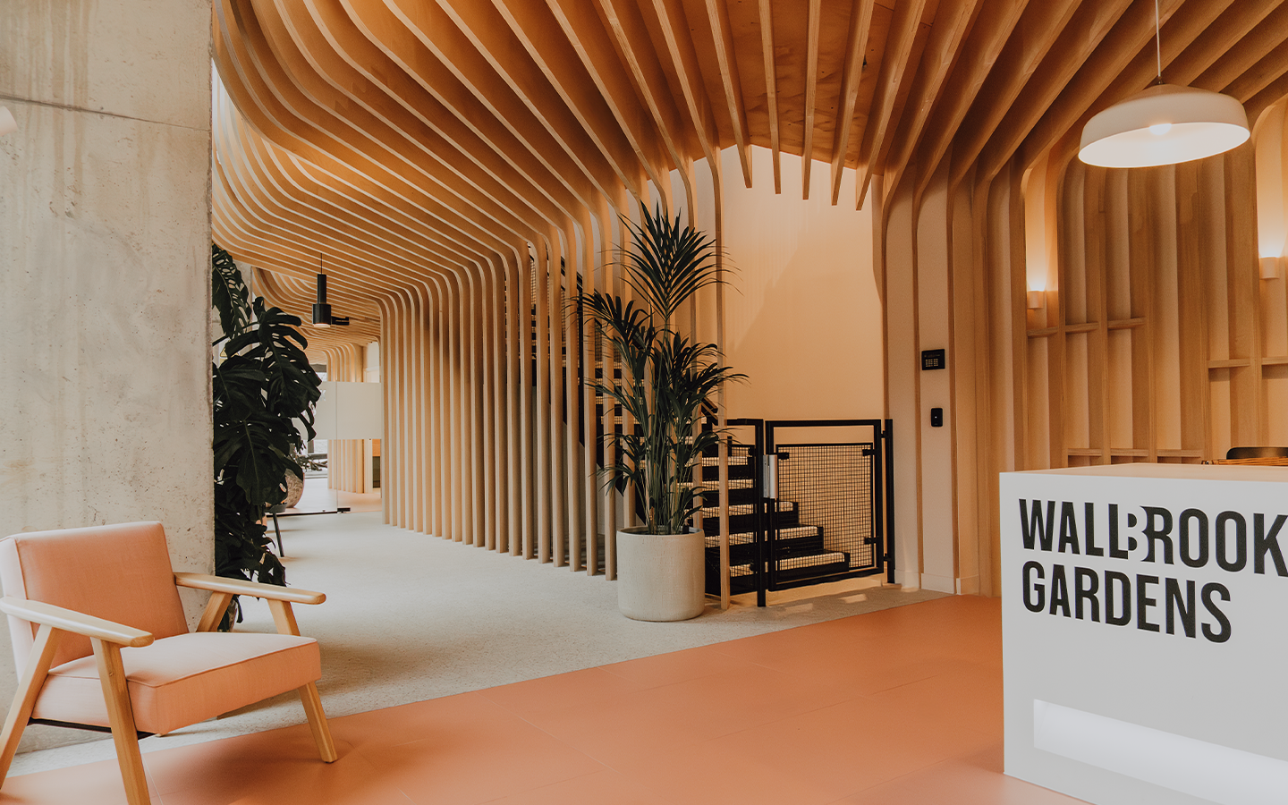Modern interior space with wooden curved ceiling beams, potted plants, a pink armchair, and a reception desk labeled 'Wallbrook Gardens'.