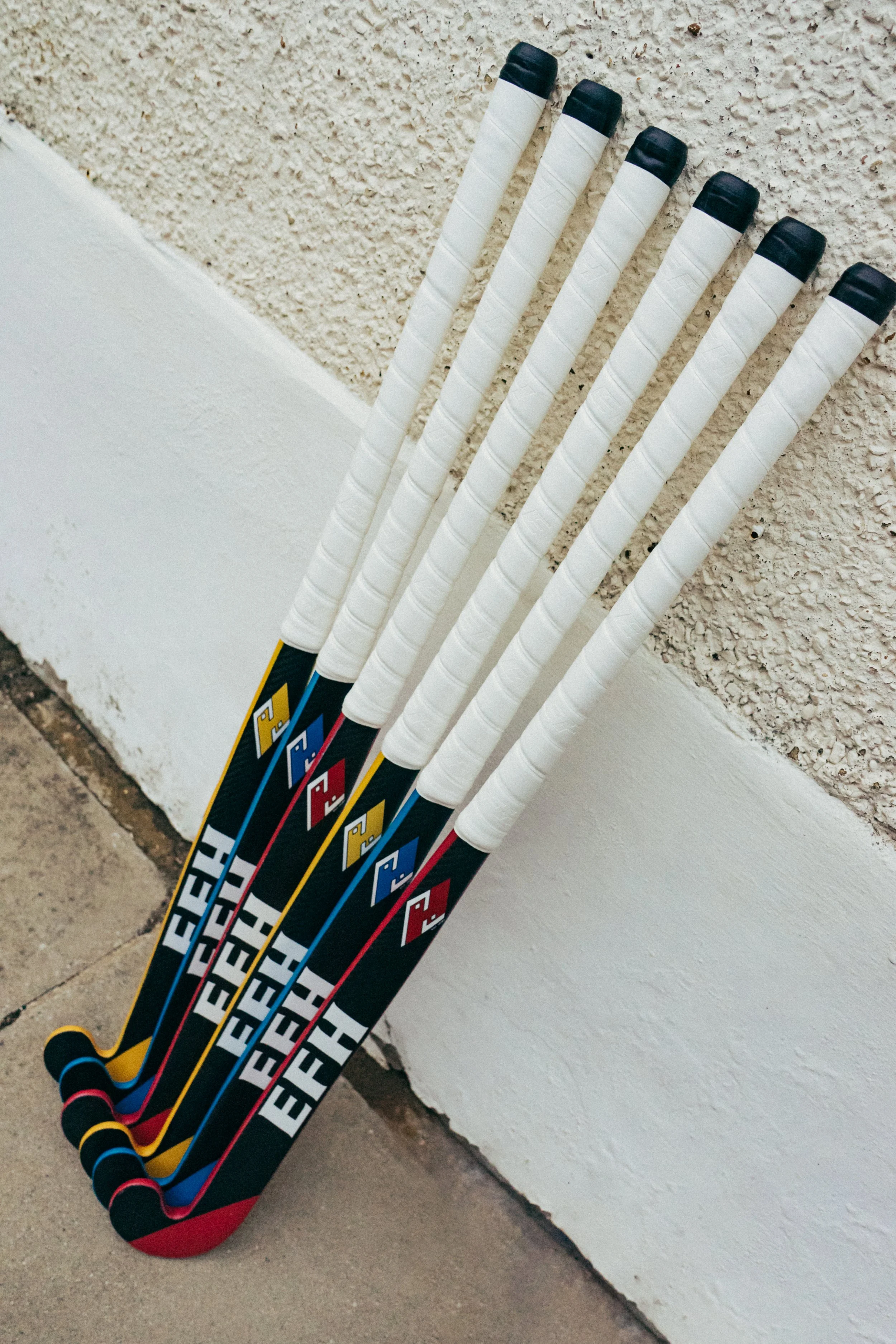 Six hockey sticks with black, white, red, yellow, and blue handles leaning against a stucco wall at an angle.