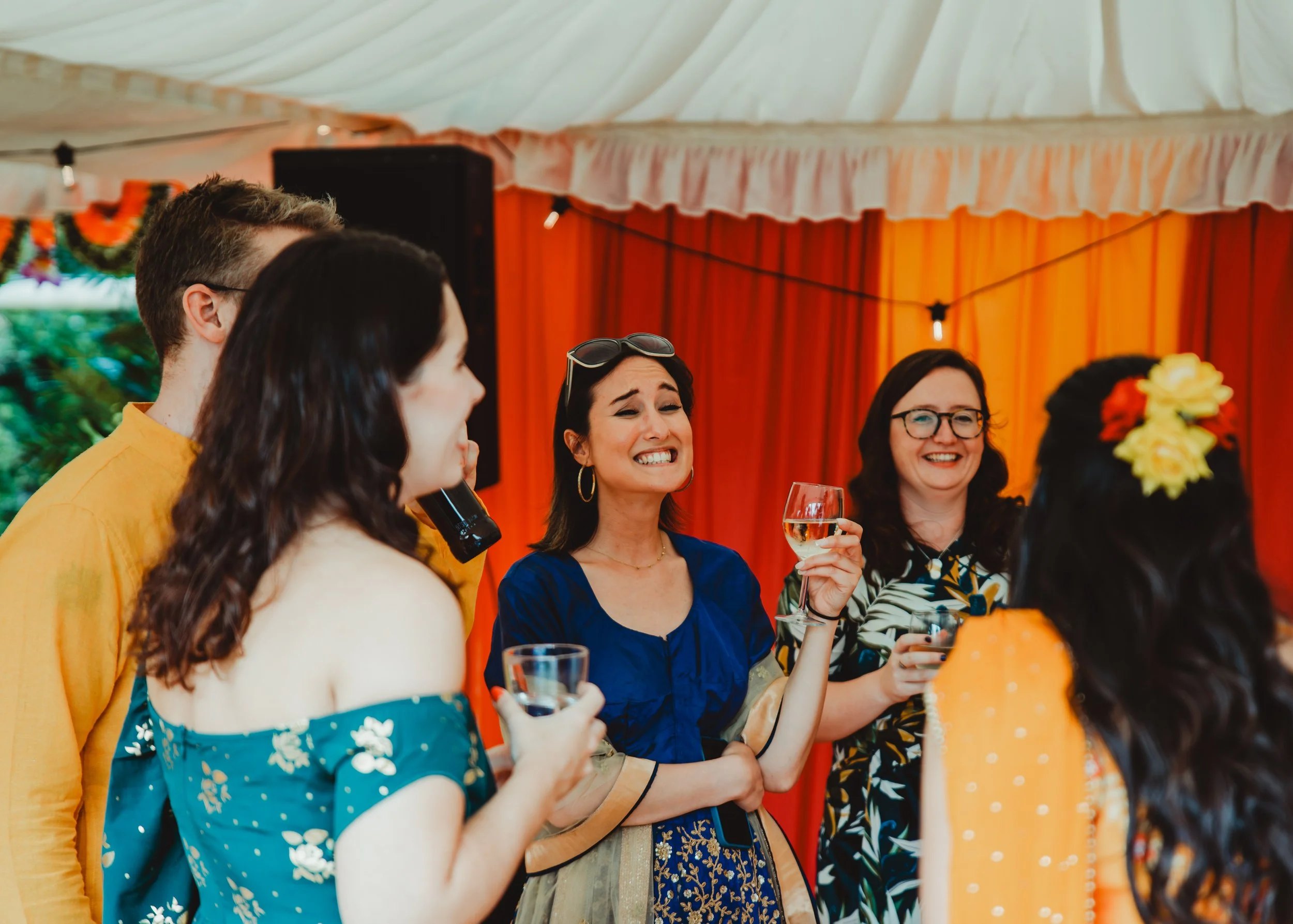 A group of women and men at a festive gathering, smiling and holding glasses of wine, with a decorated background including orange curtains and floral arrangements.