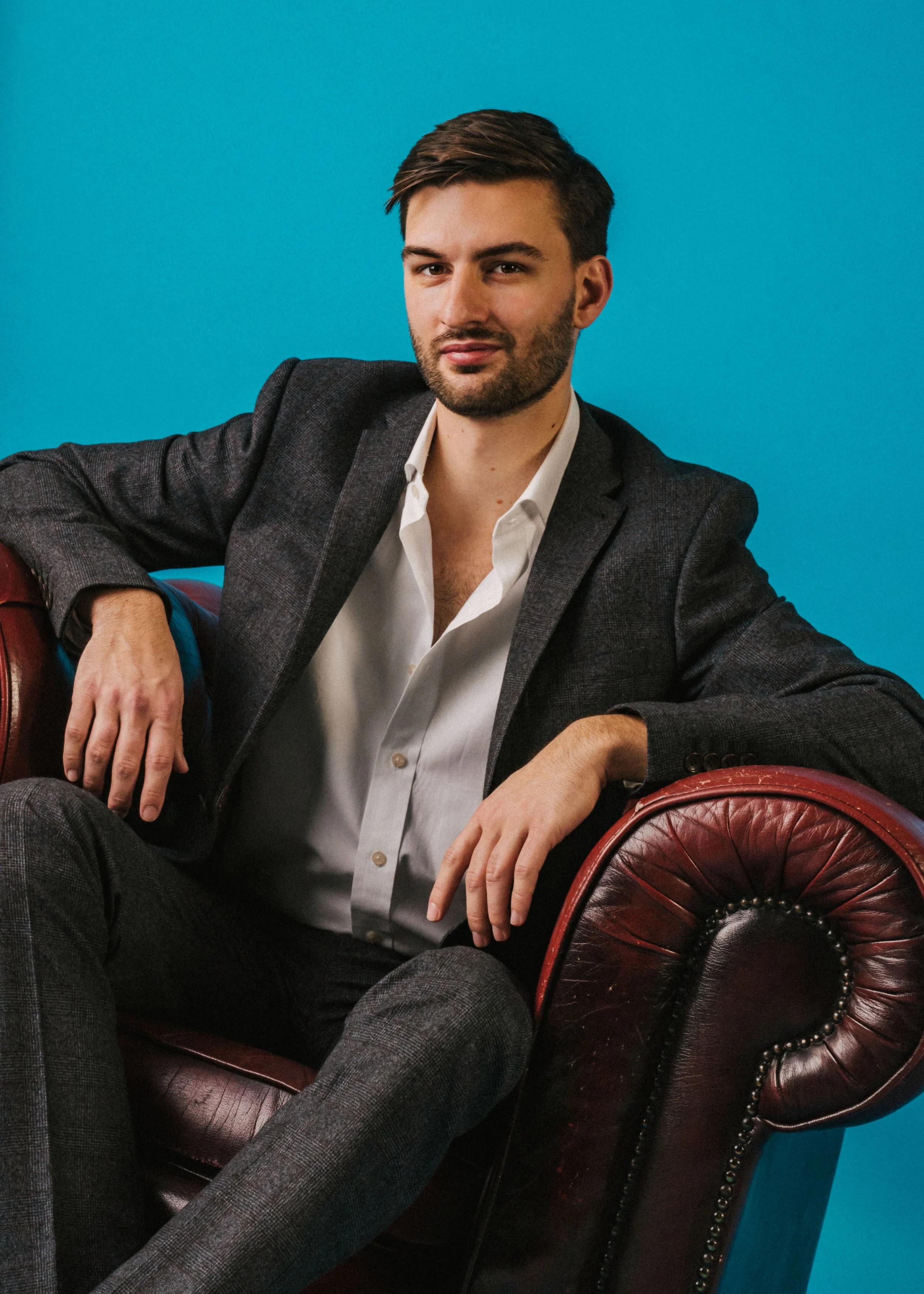 A young man with dark hair and a beard, wearing a dark blazer and a white shirt, sitting on a vintage red leather armchair with black nailhead trim against a blue background.