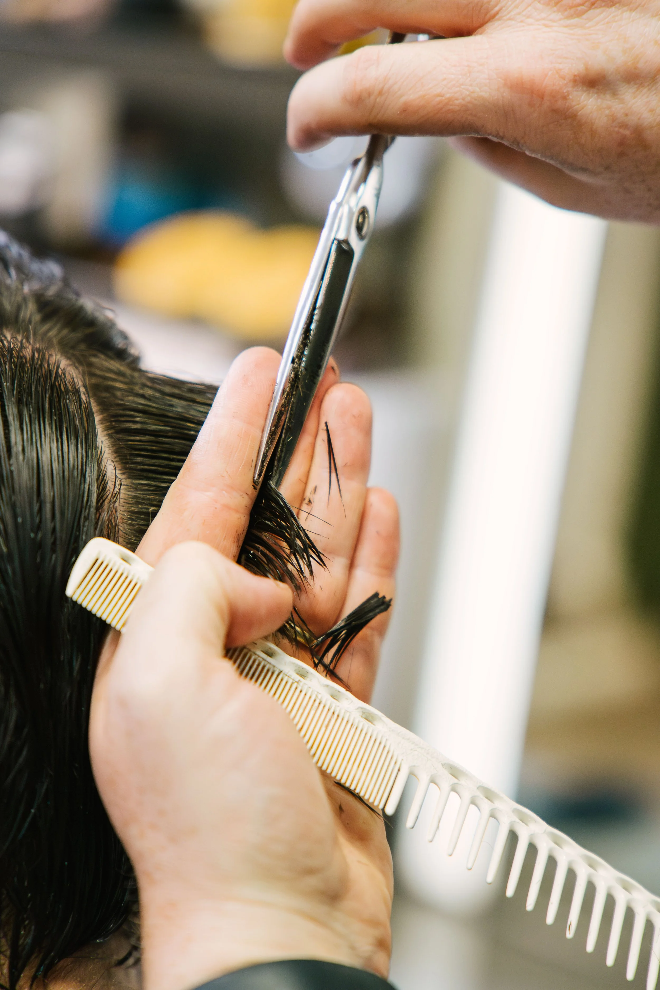 Close-up of a person cutting hair at a salon, with the hand holding scissors near the hair and a white comb underneath.
