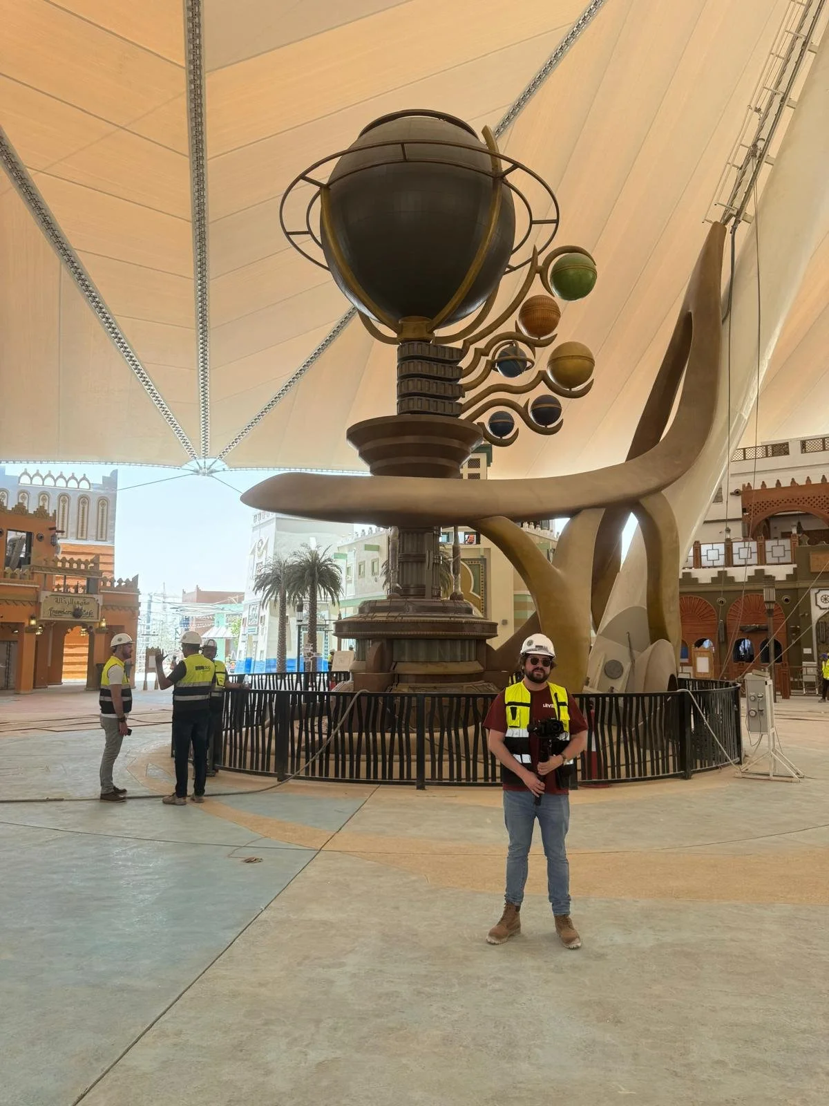 A man in a yellow safety vest and sunglasses standing in front of a large sculpture of a stylized light bulb with planetary orbits around it inside a themed indoor space. Two workers in safety vests and helmets are talking nearby, and other decorative buildings and palm trees are in the background.