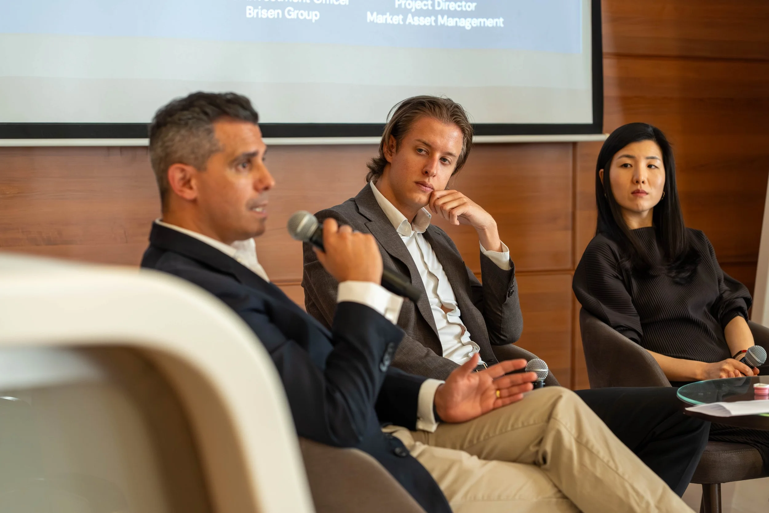 Three professionals seated on a panel during a conference or meeting, with a wooden wall behind them and a presentation slide on the wall.