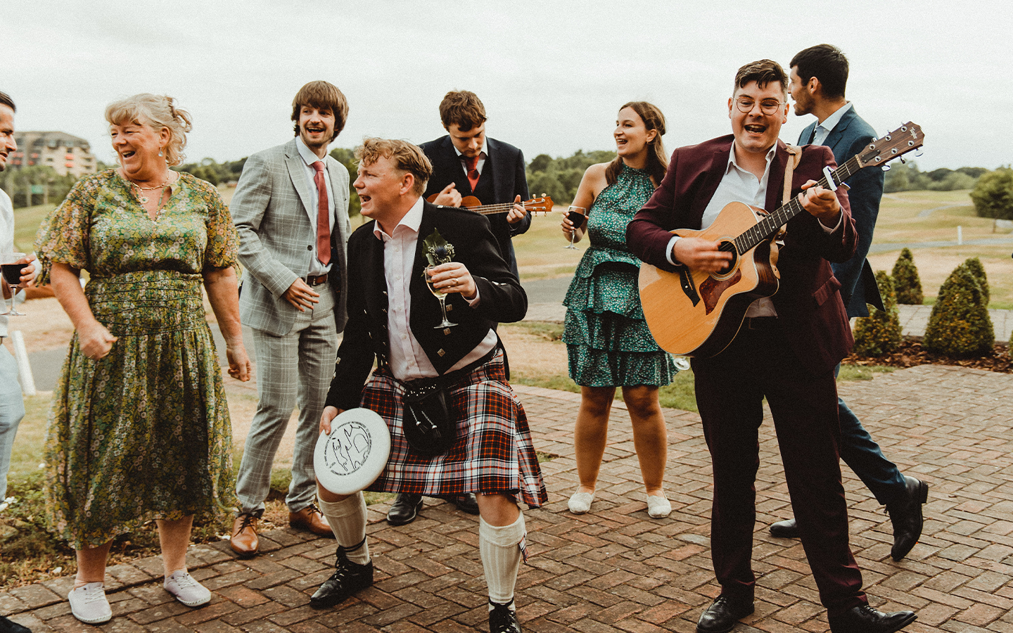 Group of people enjoying music and dancing outdoors, some playing guitars and some holding drinks, celebrating in a casual or semi-formal setting.