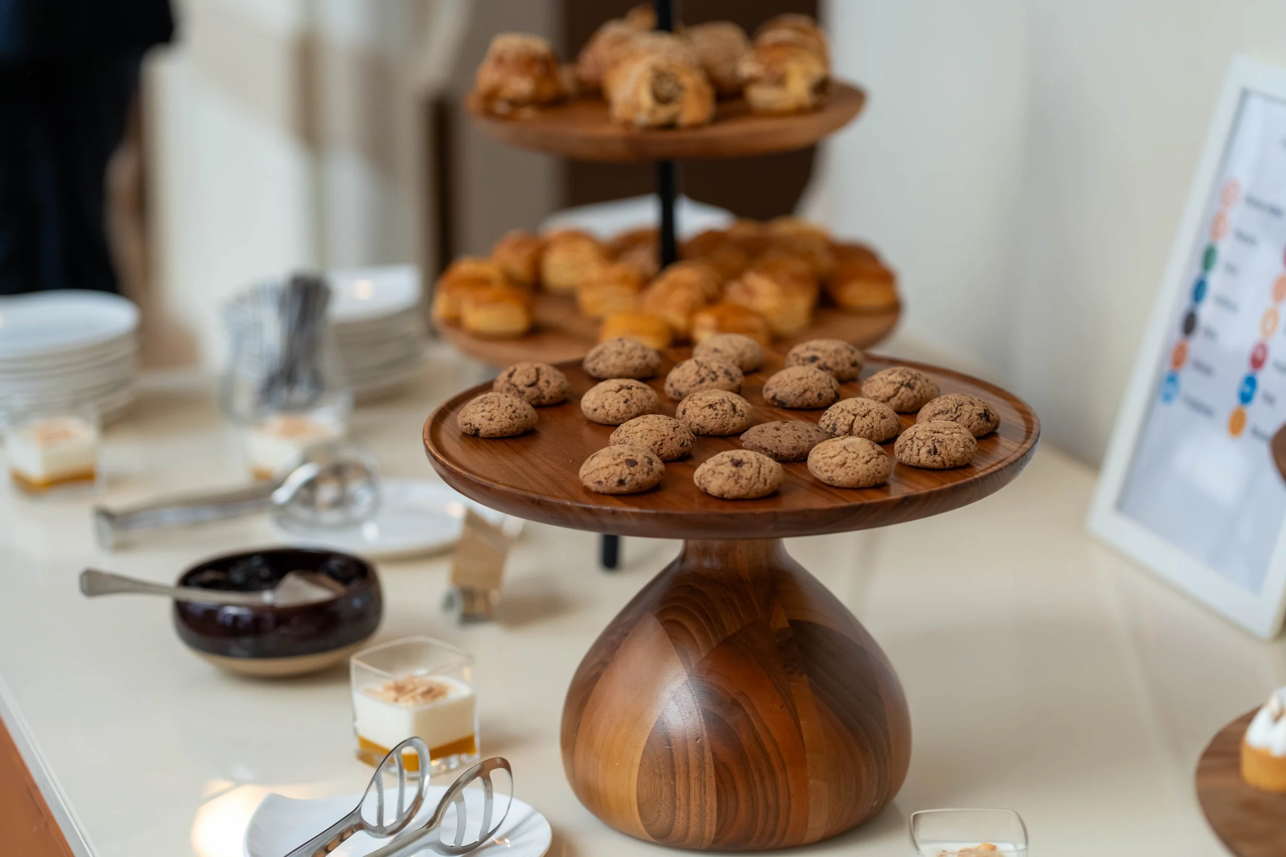 A multi-tiered wooden dessert stand displaying cookies, scones, and pastries at a table, with a blurred background of small glass cups and a menu.