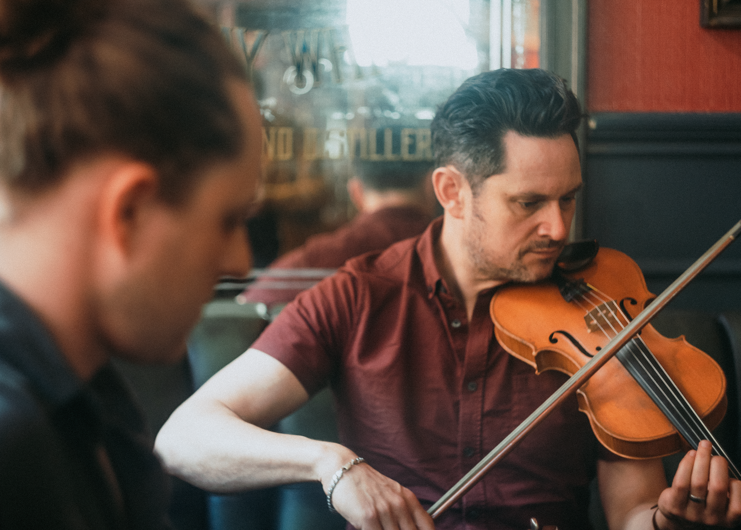 A man playing the violin, seen from a side profile with another person in the foreground, in a setting with warm lighting and a window in the background.