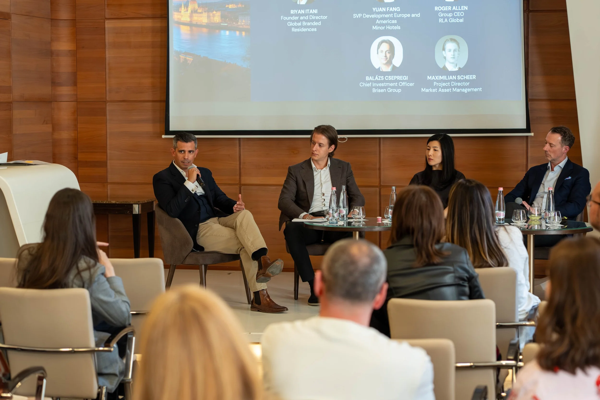 A panel of five professionals seated at a discussion table during a business conference, with a large presentation screen displaying names and titles behind them, in a wood-paneled conference room filled with an audience.