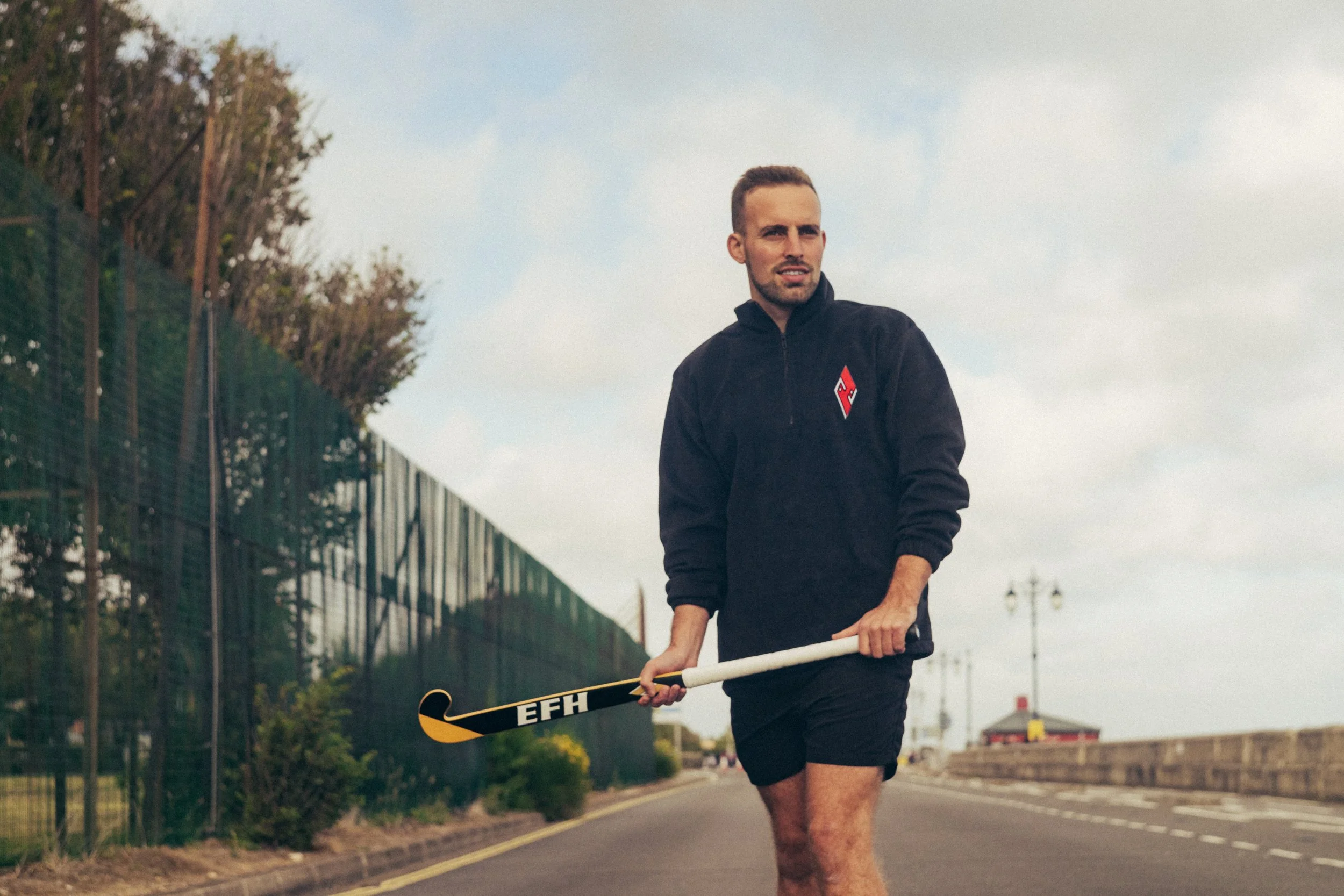 A man jogging outdoors on a pathway holding a hockey stick, wearing a black jacket with red and white logo, and black shorts. Trees and street lamps line the pathway.