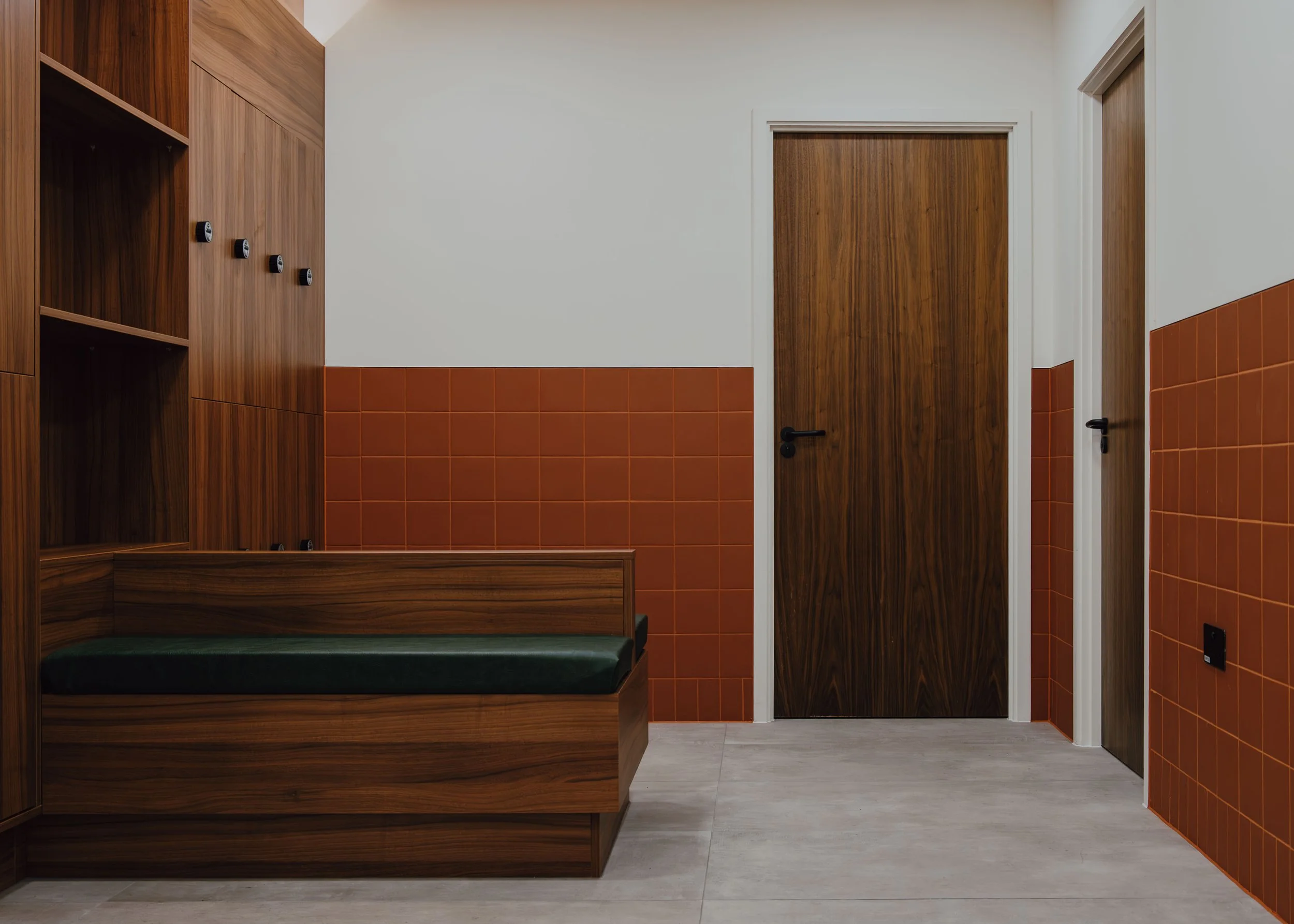 Interior of a room with wooden and orange tiled walls, two closed wooden doors, a wooden bench, and a green cushioned seating area.