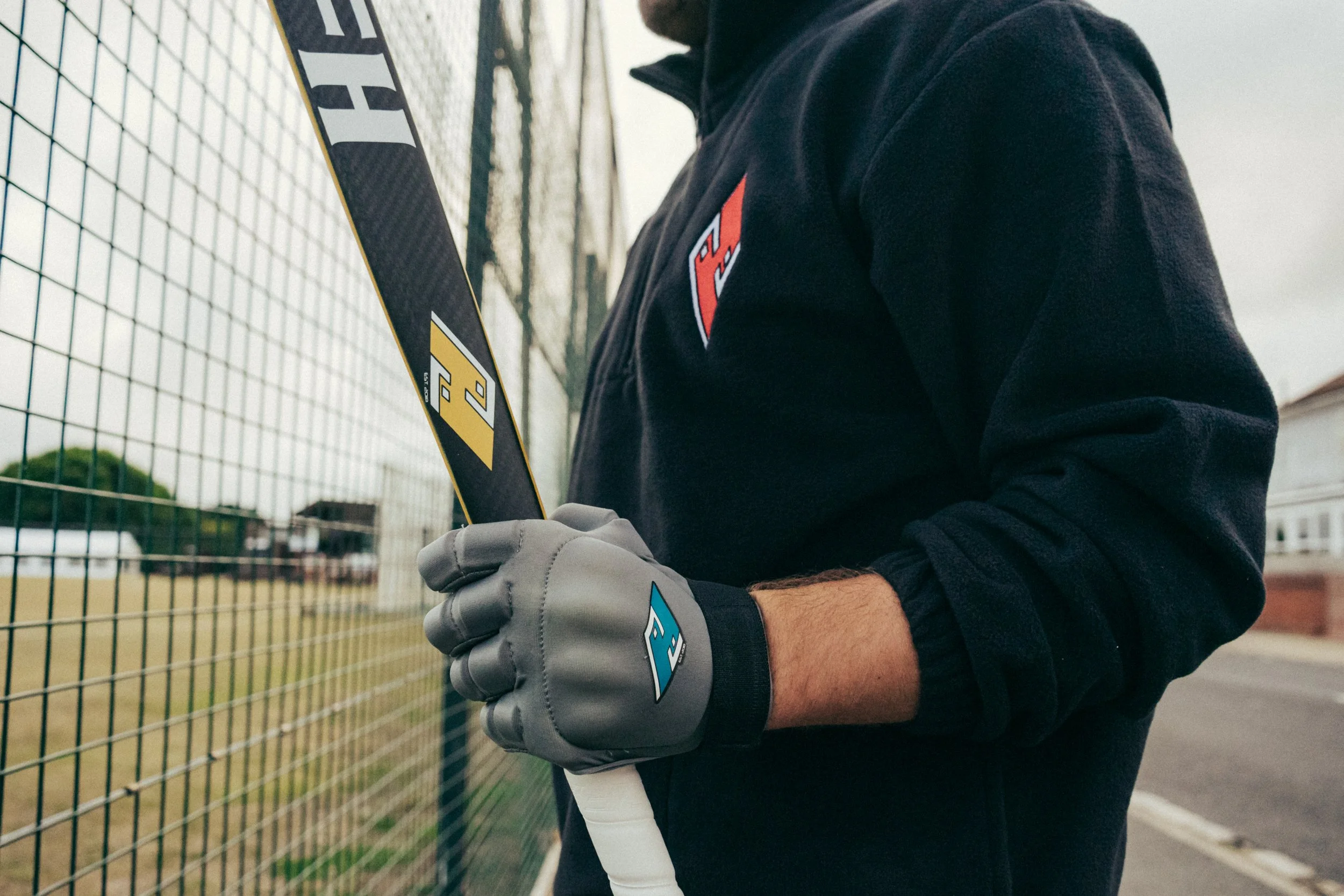 A person wearing gray gloves holding a baseball bat, standing next to a chain-link fence on an outdoor baseball field.