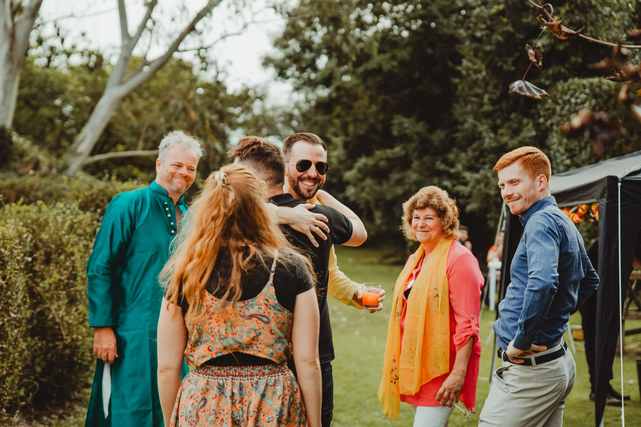 Group of people smiling and hugging at an outdoor gathering in a park, with trees and a tent in the background.