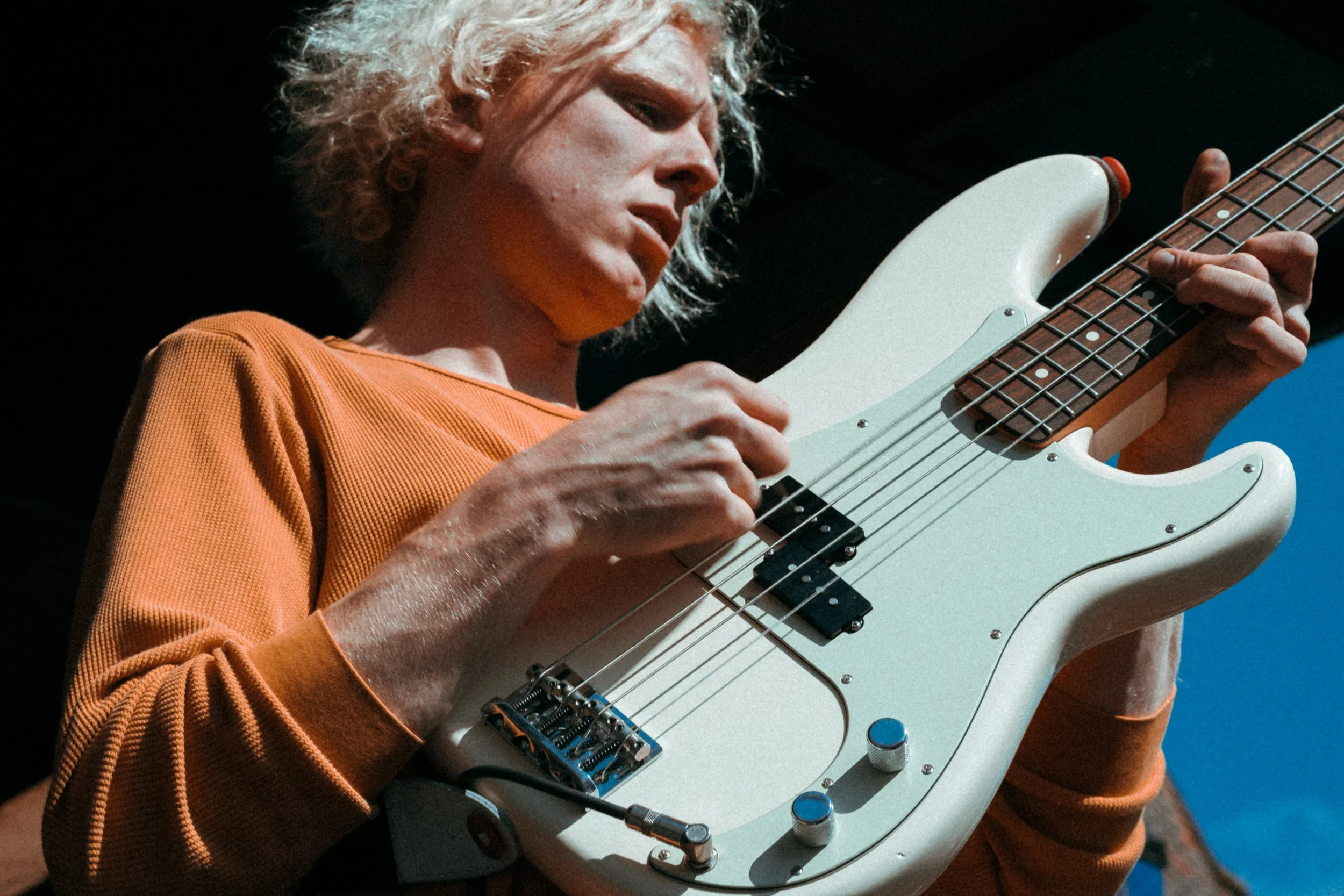 A person with curly blond hair is playing a white electric bass guitar.