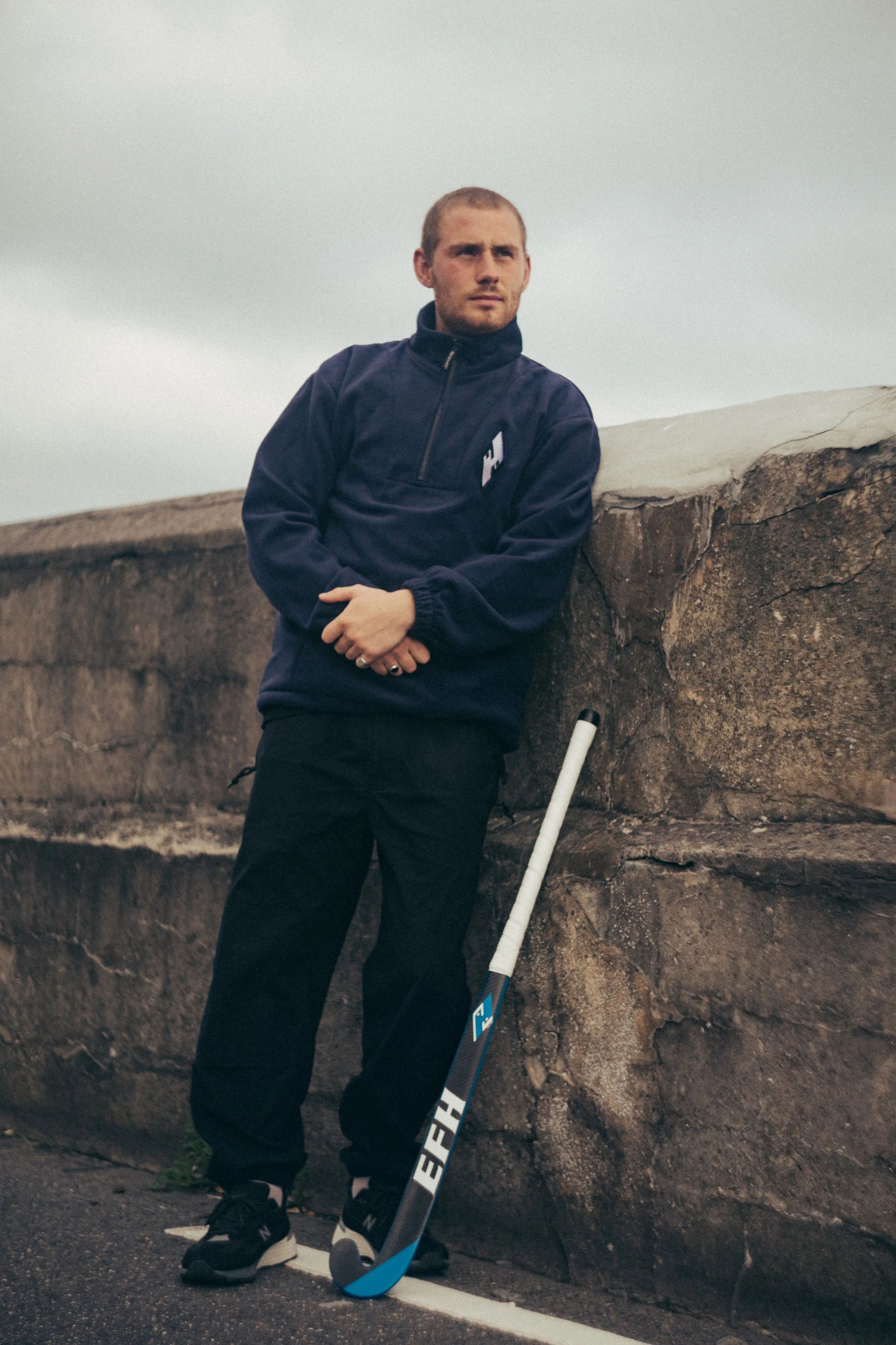 A young man standing outdoors next to a large rock wall, holding a hockey stick, looking into the distance with a serious expression. He is dressed in a navy blue quarter-zip jacket, black pants, and sneakers, with gray short hair and a light beard. 