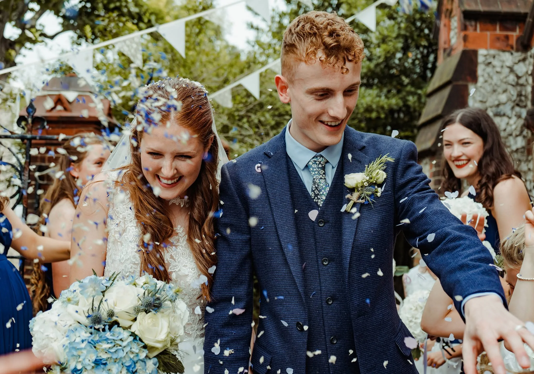 A group of people celebrating at an outdoor wedding, with a bride and groom smiling as they walk through falling flower petals, surrounded by friends and family, under string lights and bunting.