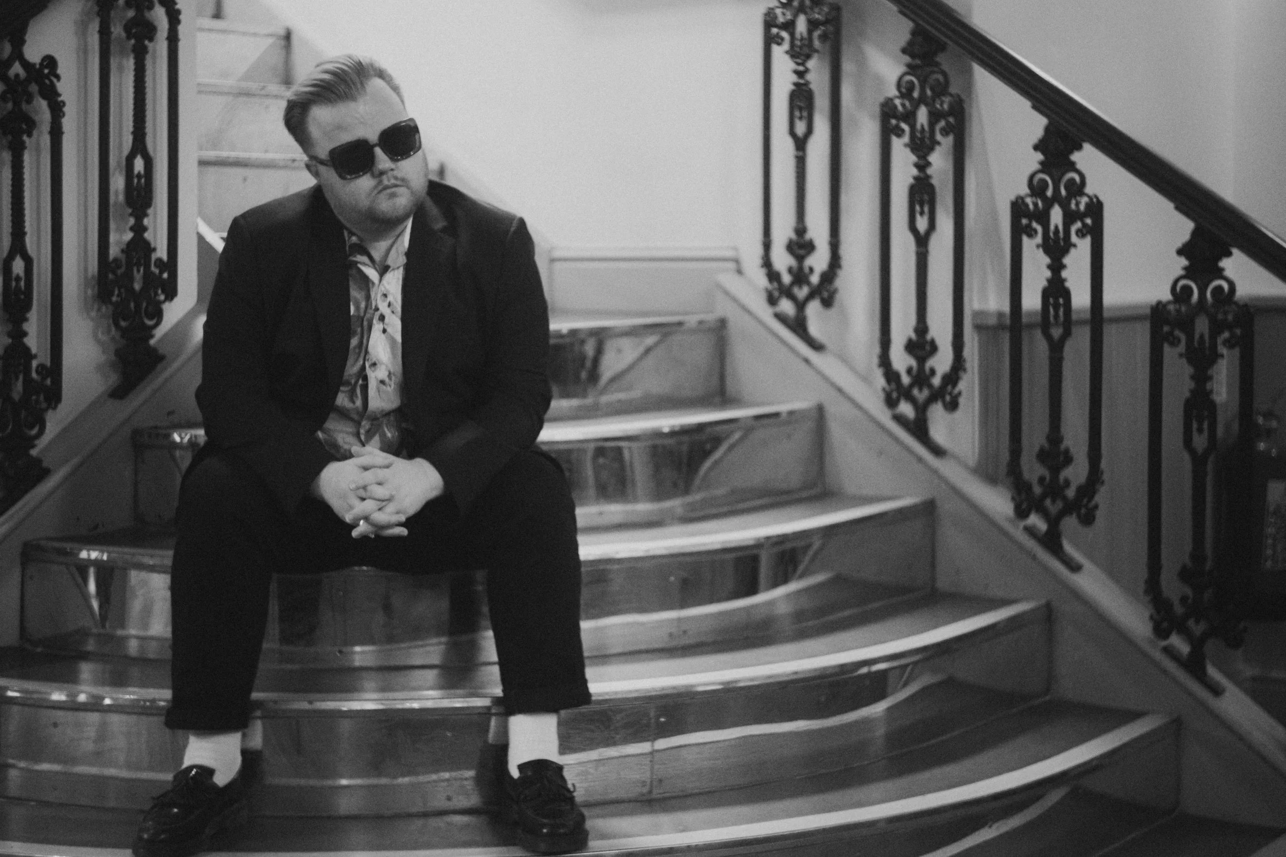 A man with light-colored hair, sunglasses, and a beard sitting on a staircase with ornate wrought iron railings. He is wearing a dark jacket, patterned shirt, black pants, and dark shoes, with his hands clasped in front of him. The staircase has curv