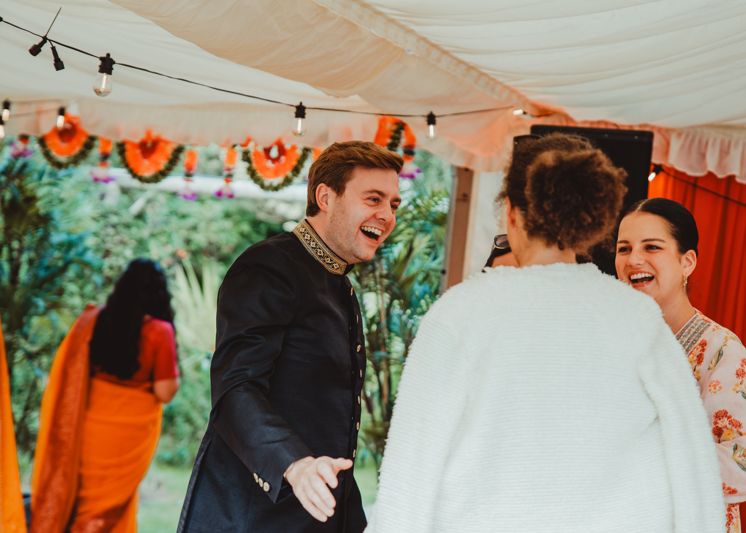 A man and woman smiling and talking, holding hands at an outdoor event decorated with orange and green garlands and string lights under a canopy.