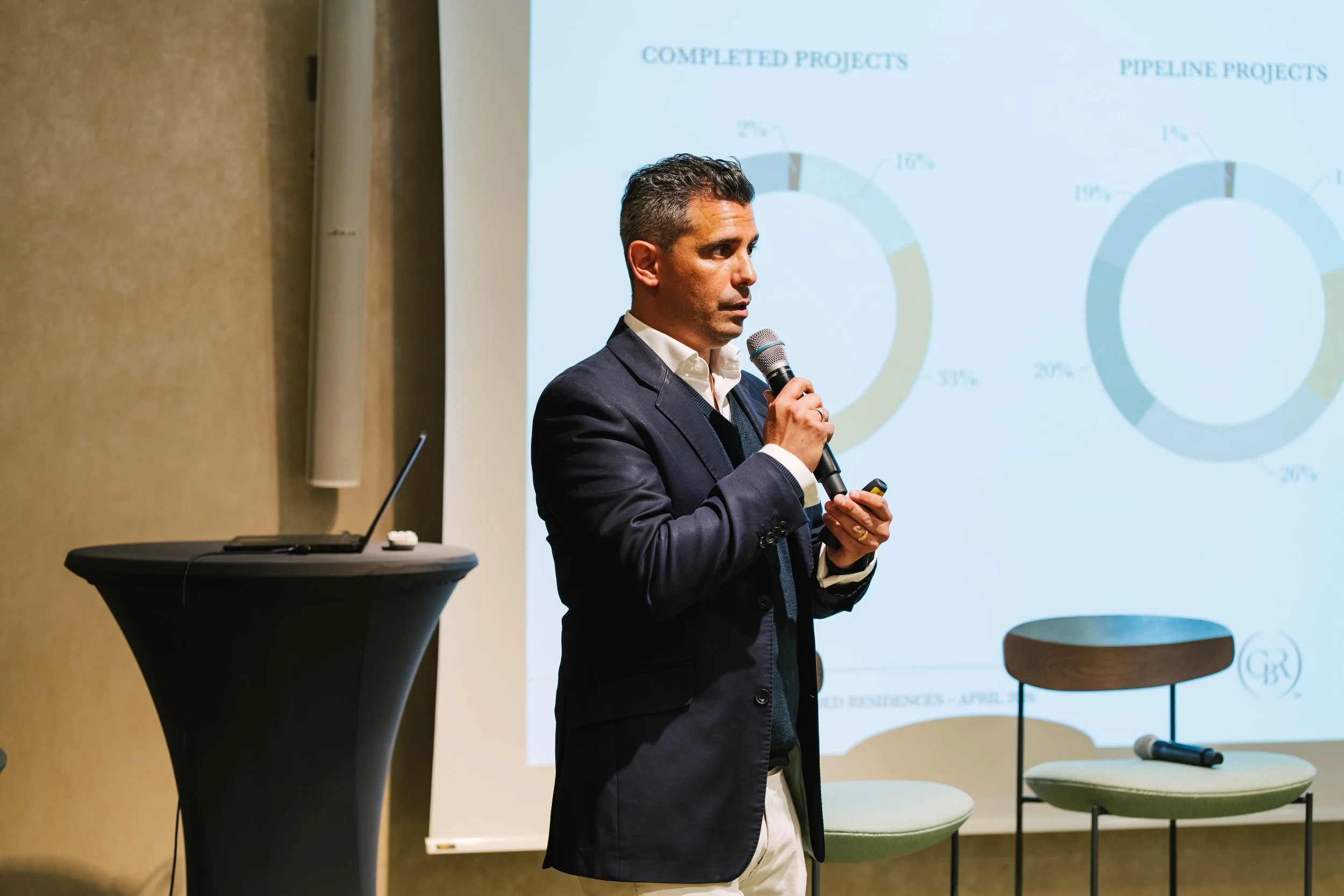 A man in a suit holding a microphone and a presentation remote, standing in front of a projection screen with pie chart graphics, in a conference room setting.