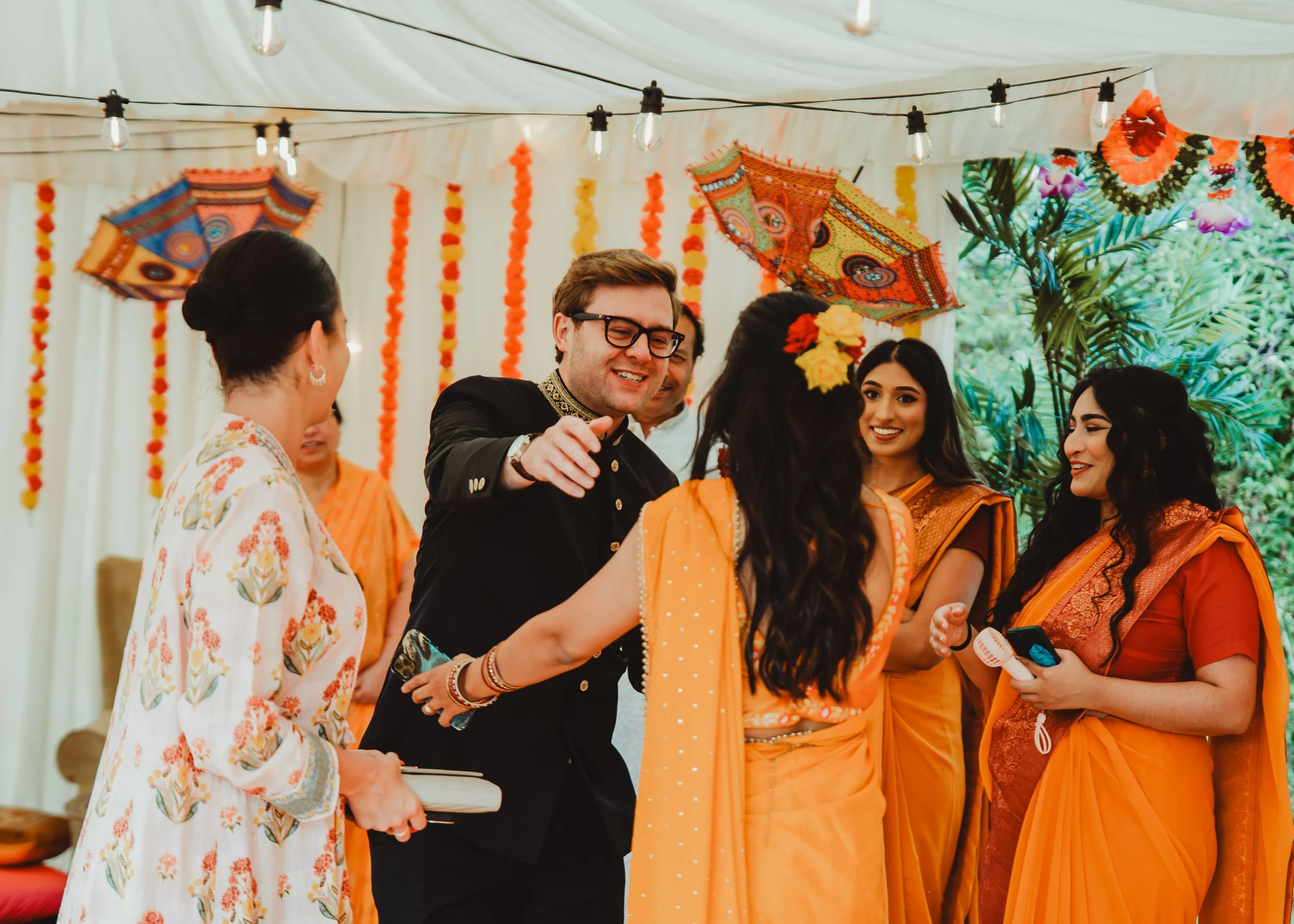 People gathered at an outdoor event decorated with string lights, colorful flowers, and hanging lanterns. Some women are dressed in traditional Indian sarees, and a man in a black suit is smiling and interacting with others.