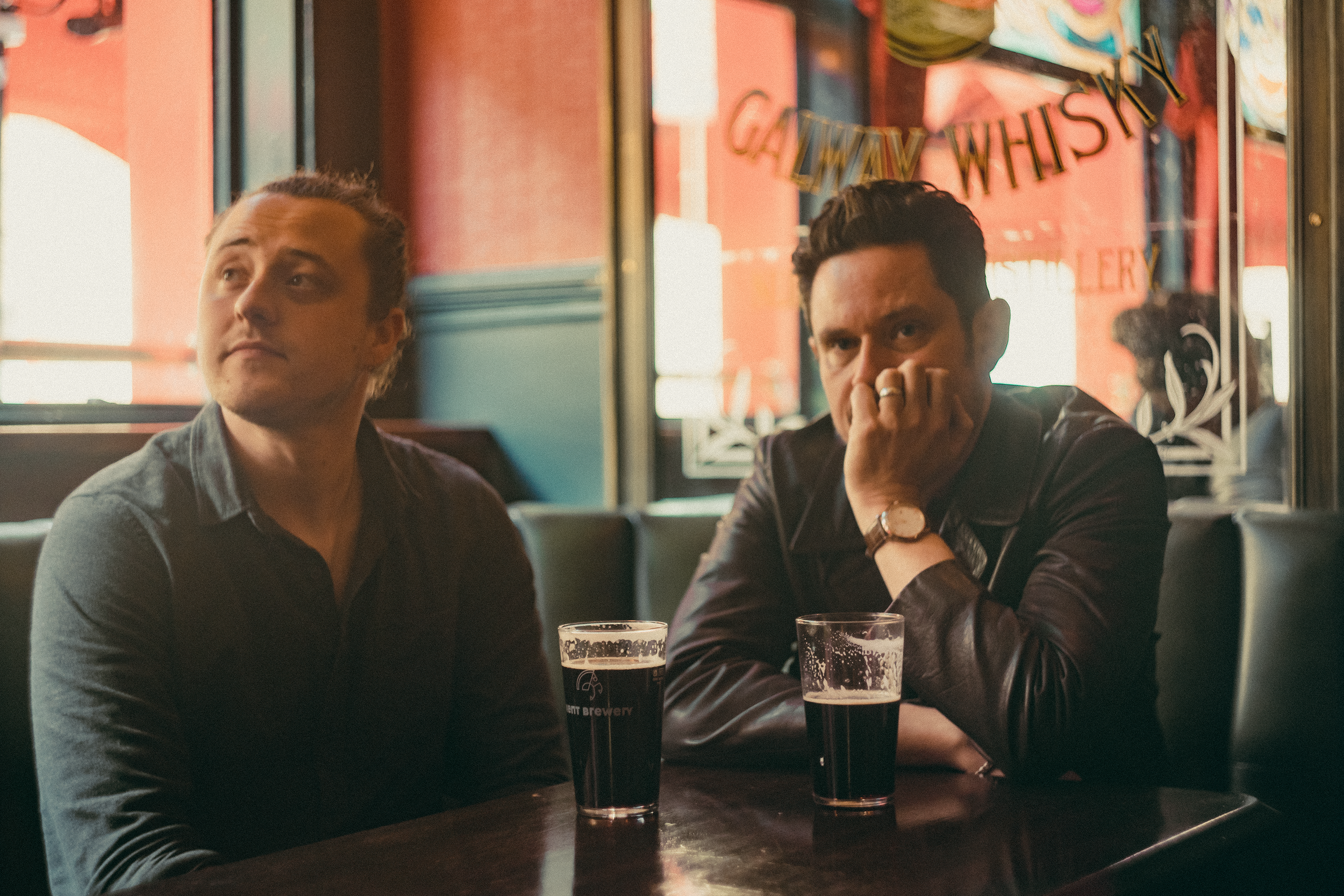 Two men sitting at a bar or restaurant table with glasses of dark beer in front of them. One man on the left has long hair tied back, and the man on the right has short dark hair and is resting his chin on his hand. They are inside with large windows