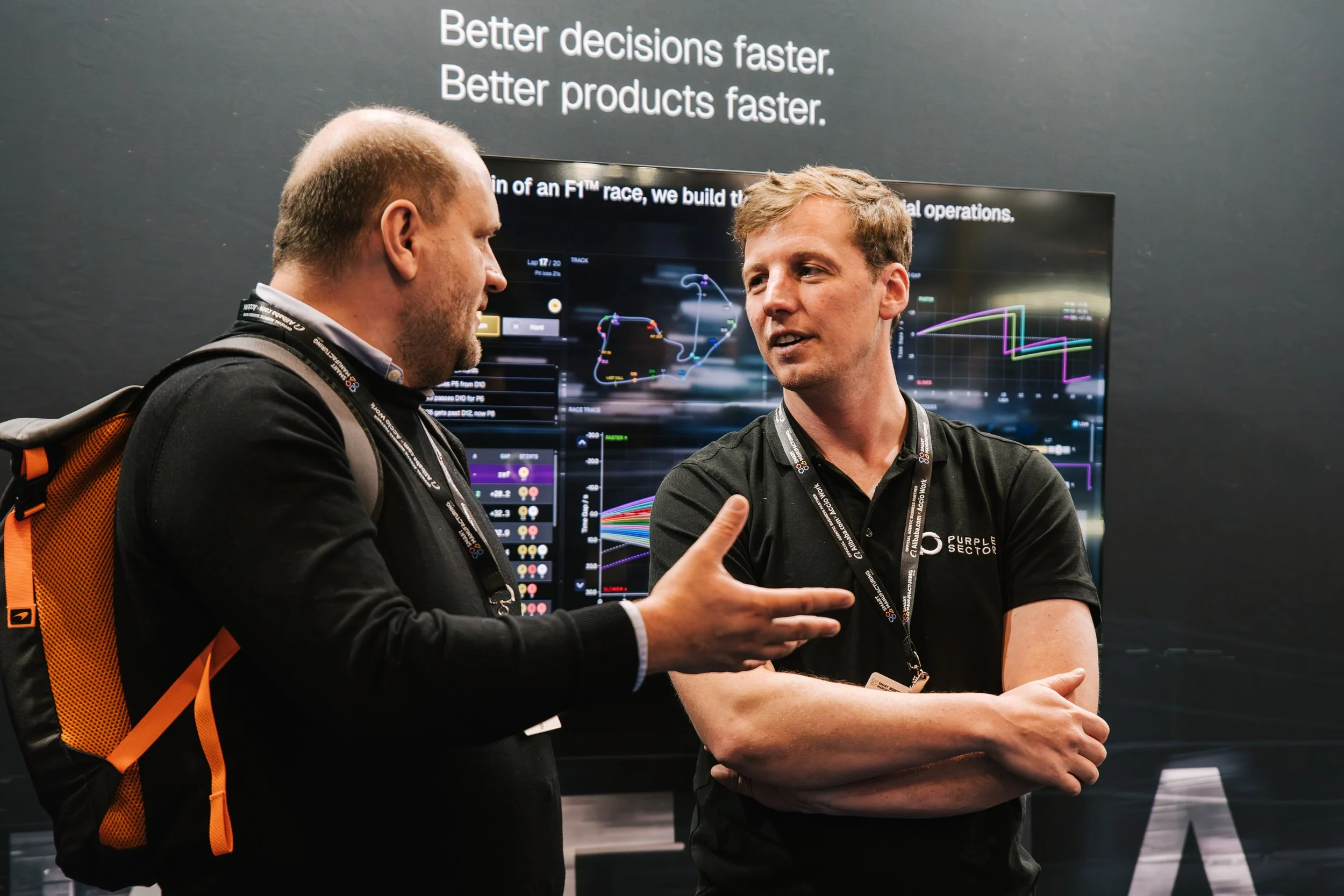 Two men engaged in conversation at a conference or tech event, with a large display screen behind them showing graphs and data related to racing or technology, and a black wall with white text saying, 'Better decisions faster. Better products faster.