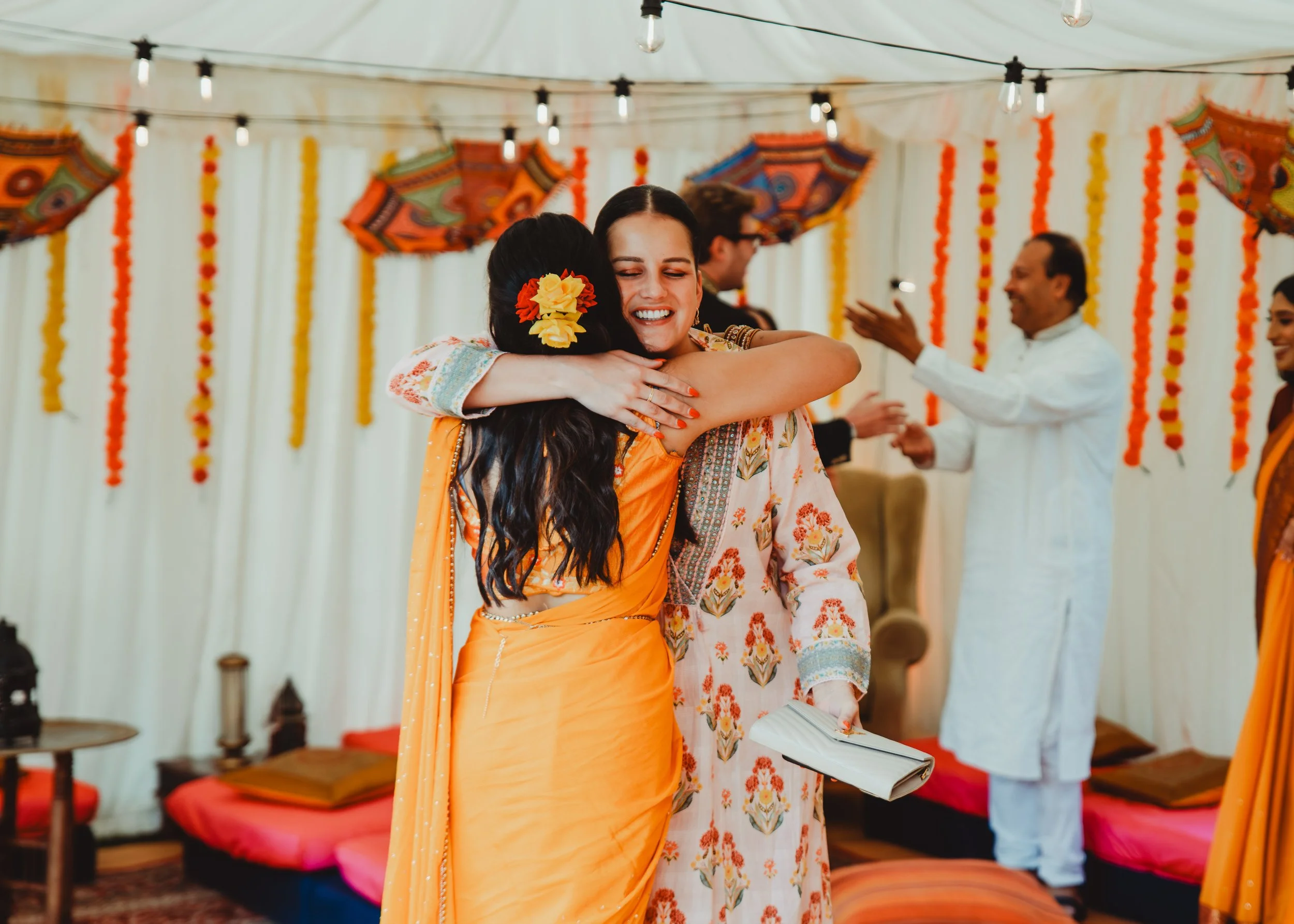 Two women in colorful traditional attire hugging at an Indian celebration, with a decorated backdrop and other guests in the background.