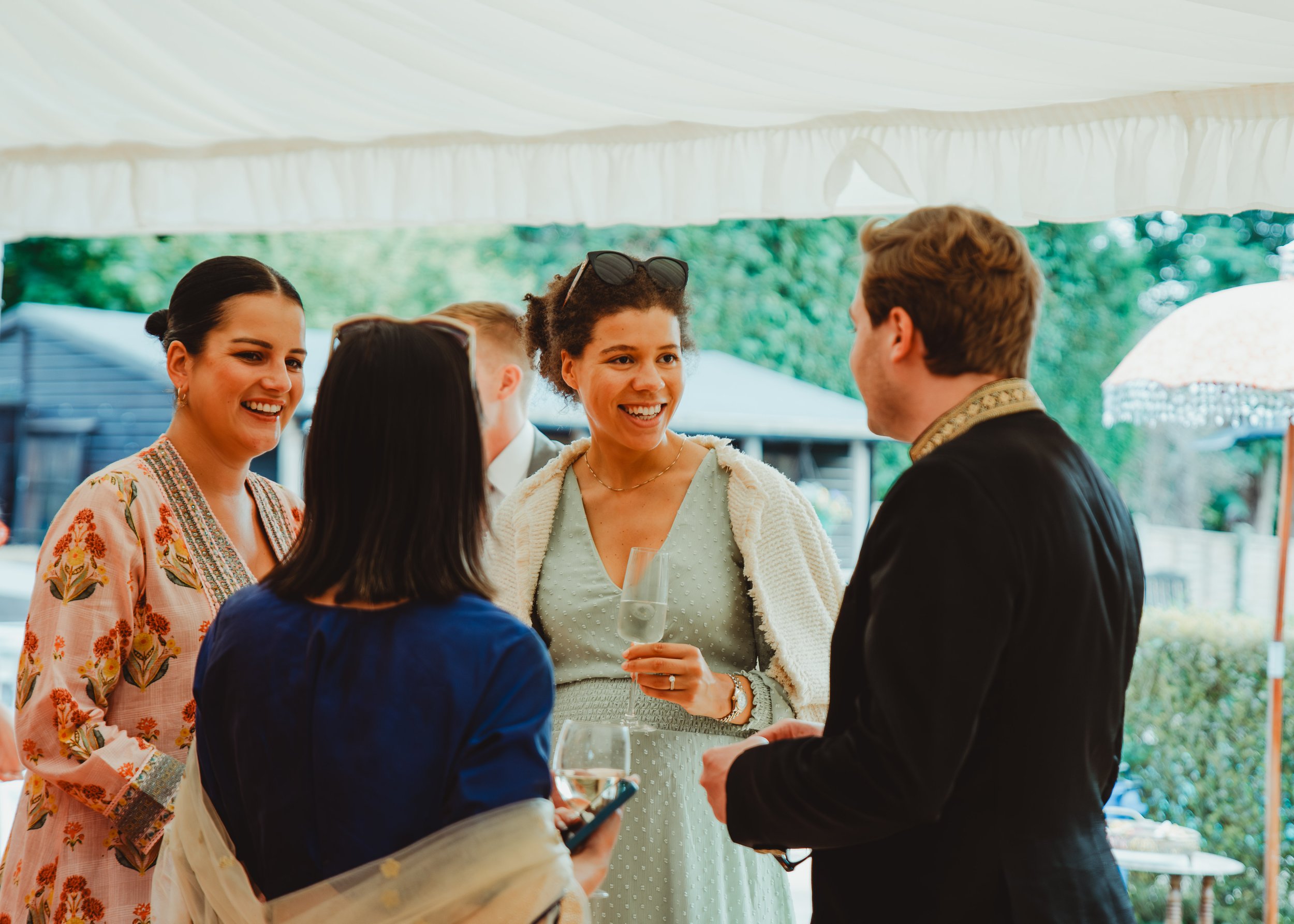 A group of five people socializing outdoors under a canopy, with one person holding a glass of wine.