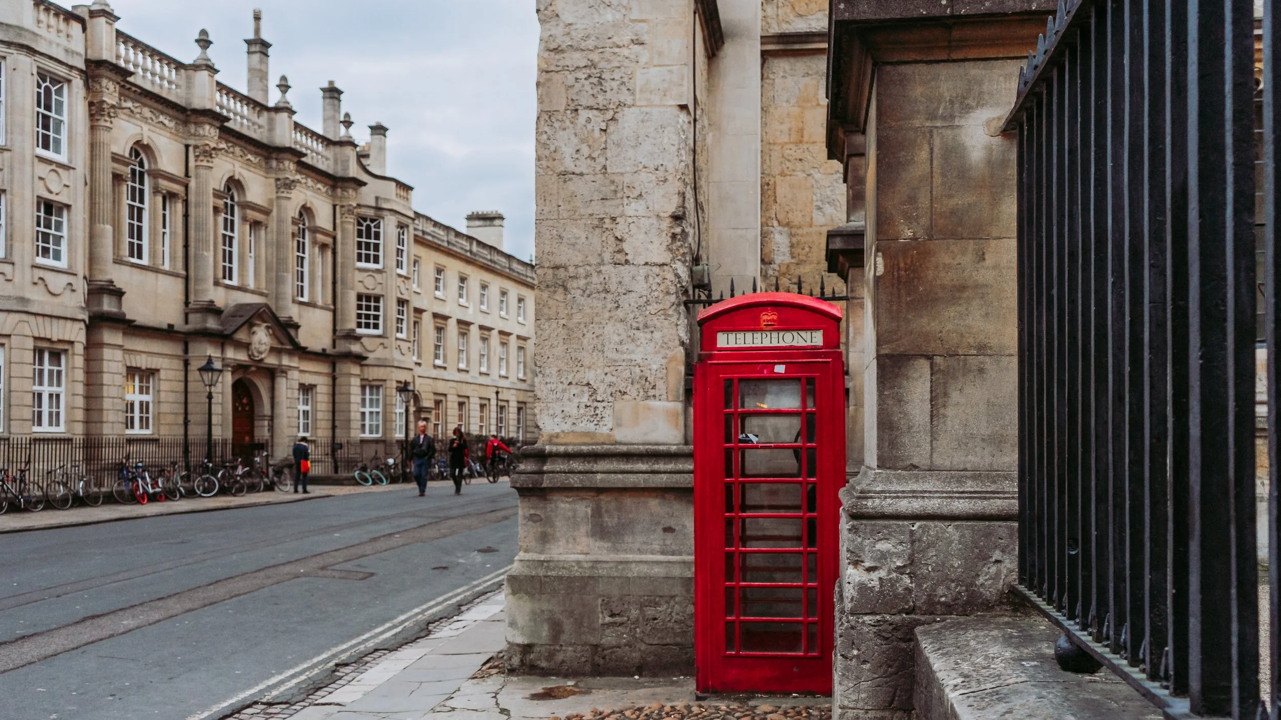 Red telephone booth on a city street with historic buildings, bicycles, and pedestrians in the background.
