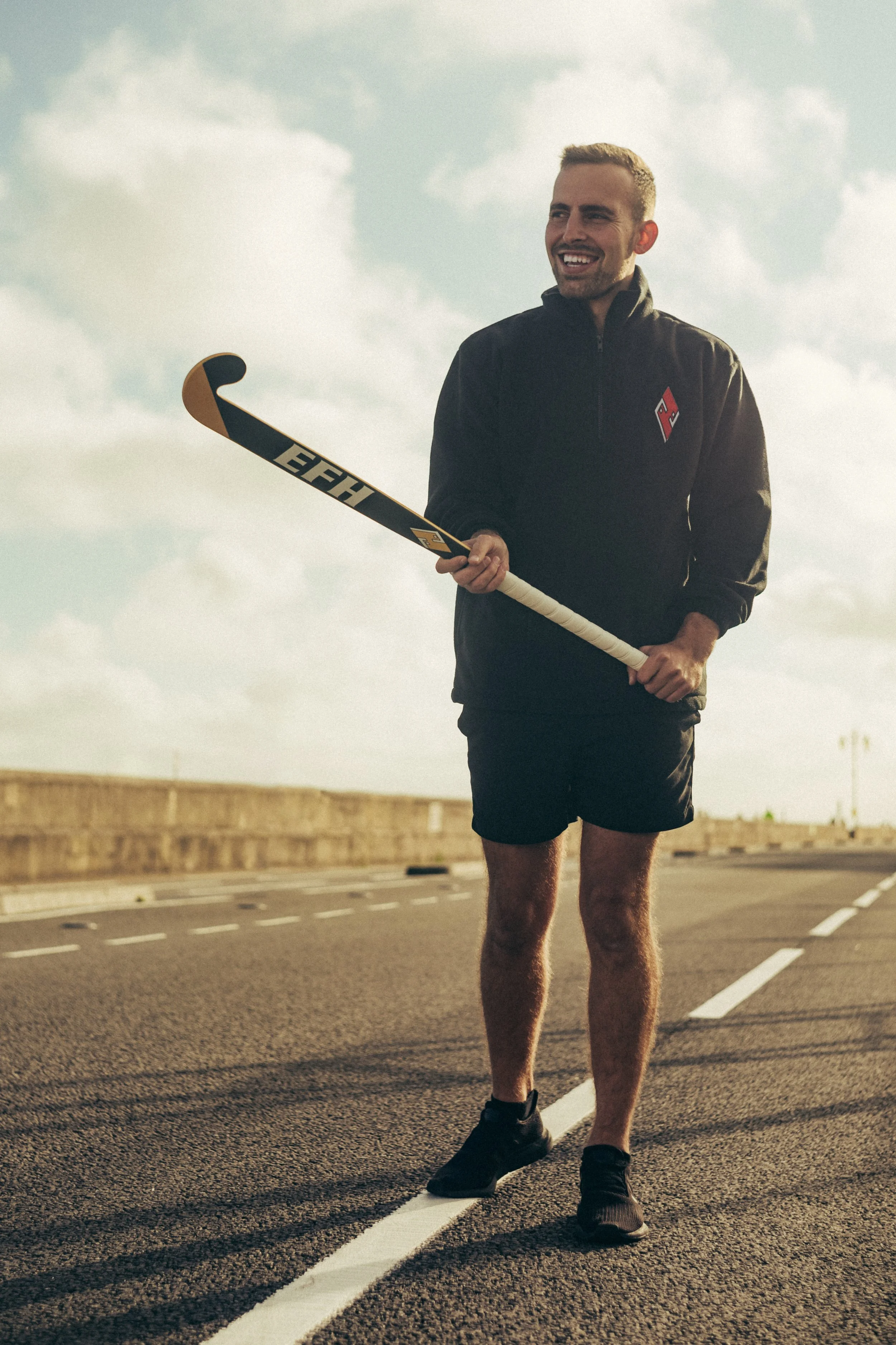 A man standing on a road holding a hockey stick, wearing a black athletic jacket and shorts, with a cloudy sky in the background.