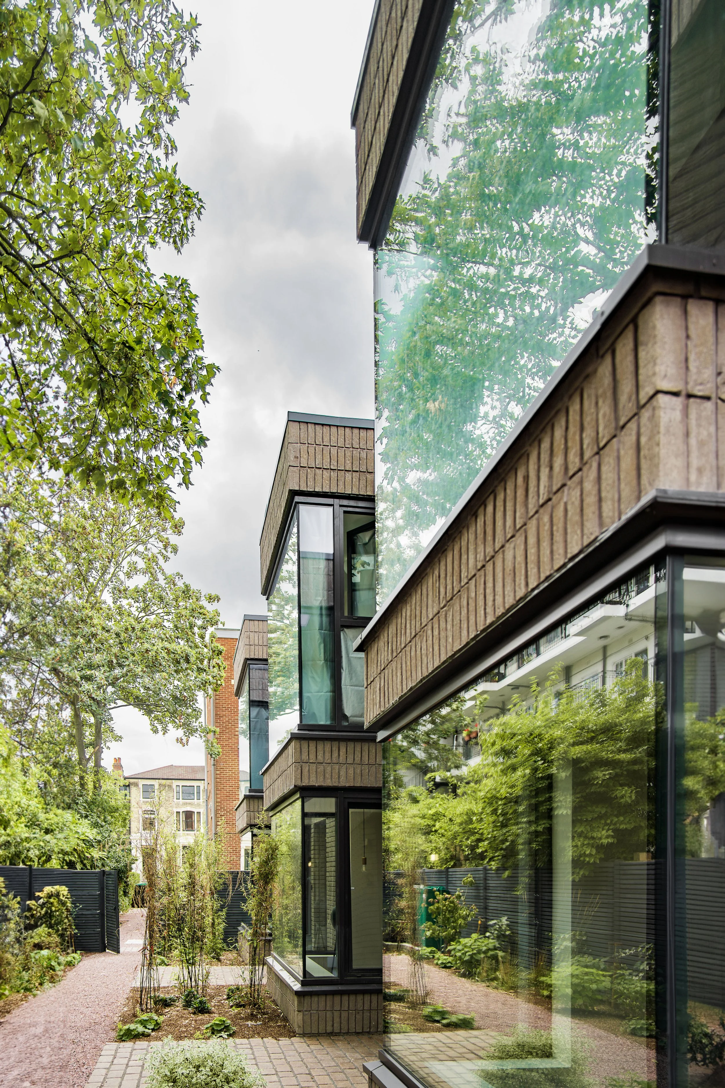 Reflective glass window on a modern multi-story building with brick facade, showing a garden with trees and plants in the foreground, and city buildings in the background, under a cloudy sky.