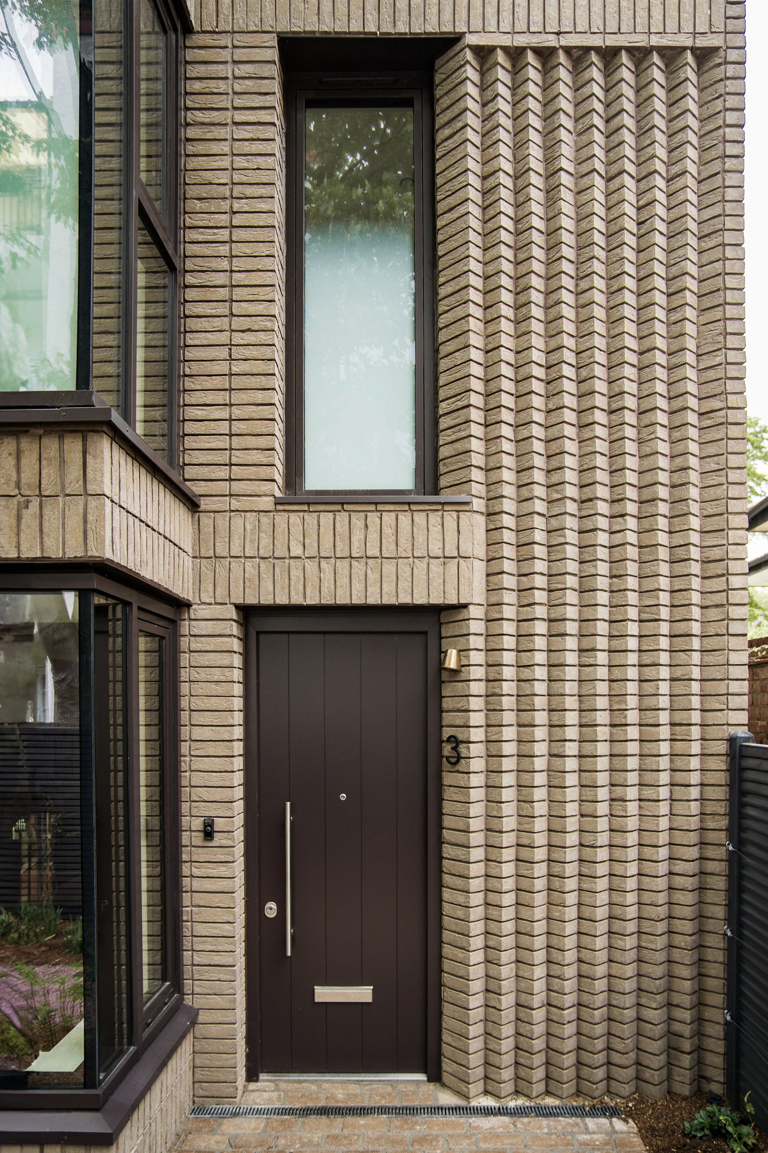 Modern brown brick building entrance with a black door, a small window above, and the number 3 to the right of the door.