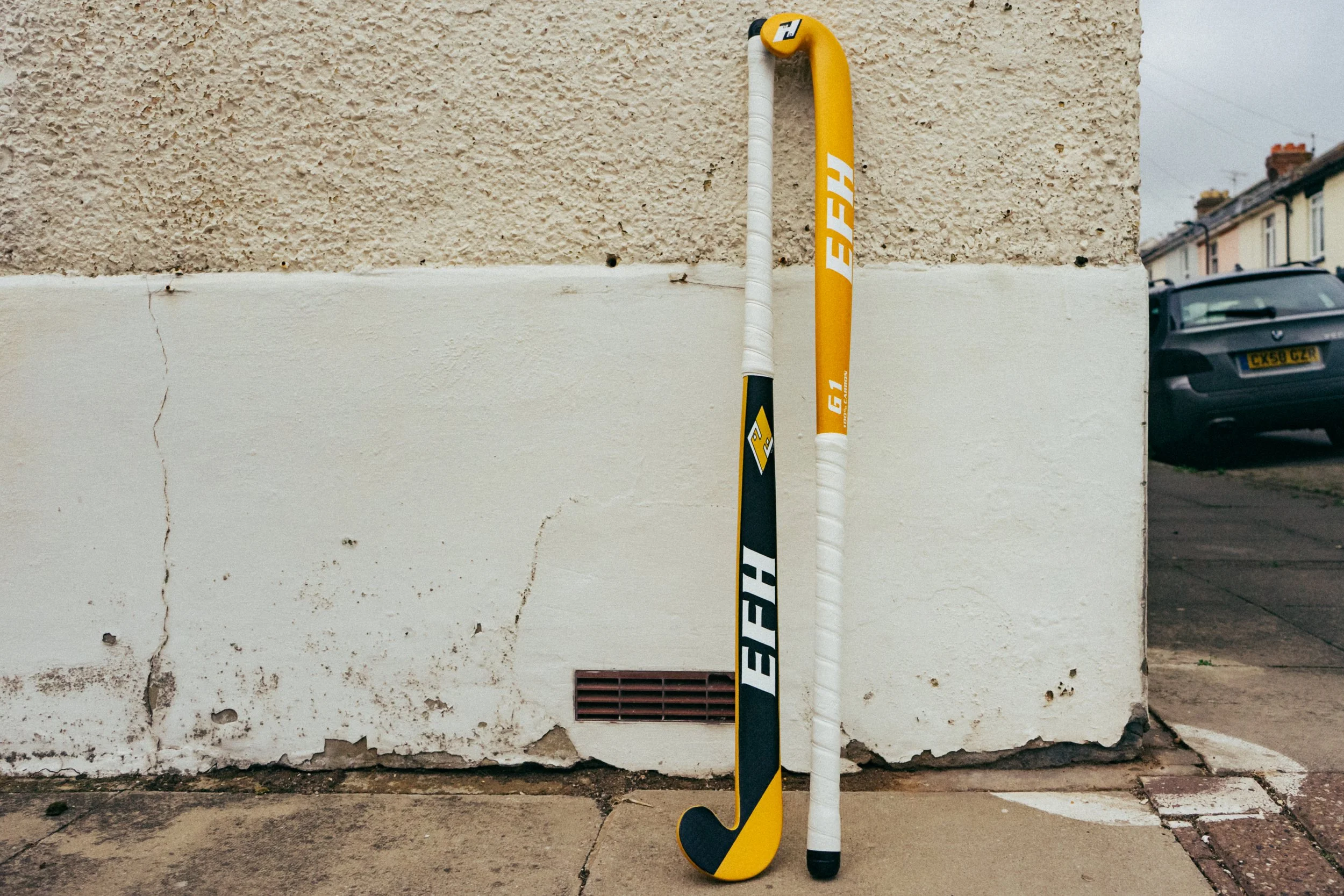 Two hockey sticks, one yellow and black, one white and black, are positioned upright against a textured beige wall with cracks. A black car is parked on the street in the background.