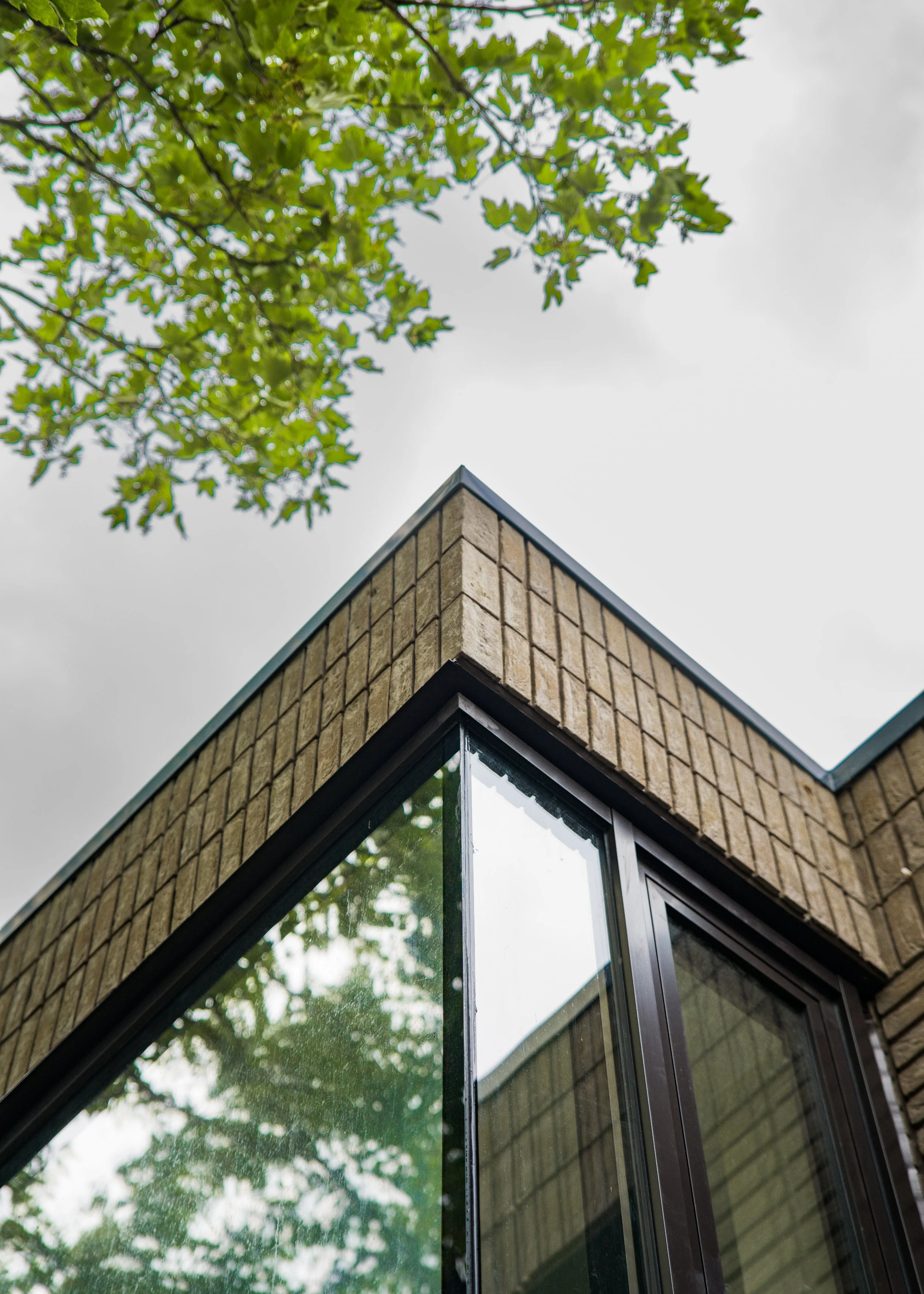 The image shows the corner of a modern building with a brick facade, large glass windows, and a sloped roof. There are green leaves from a tree in the background against a cloudy sky.