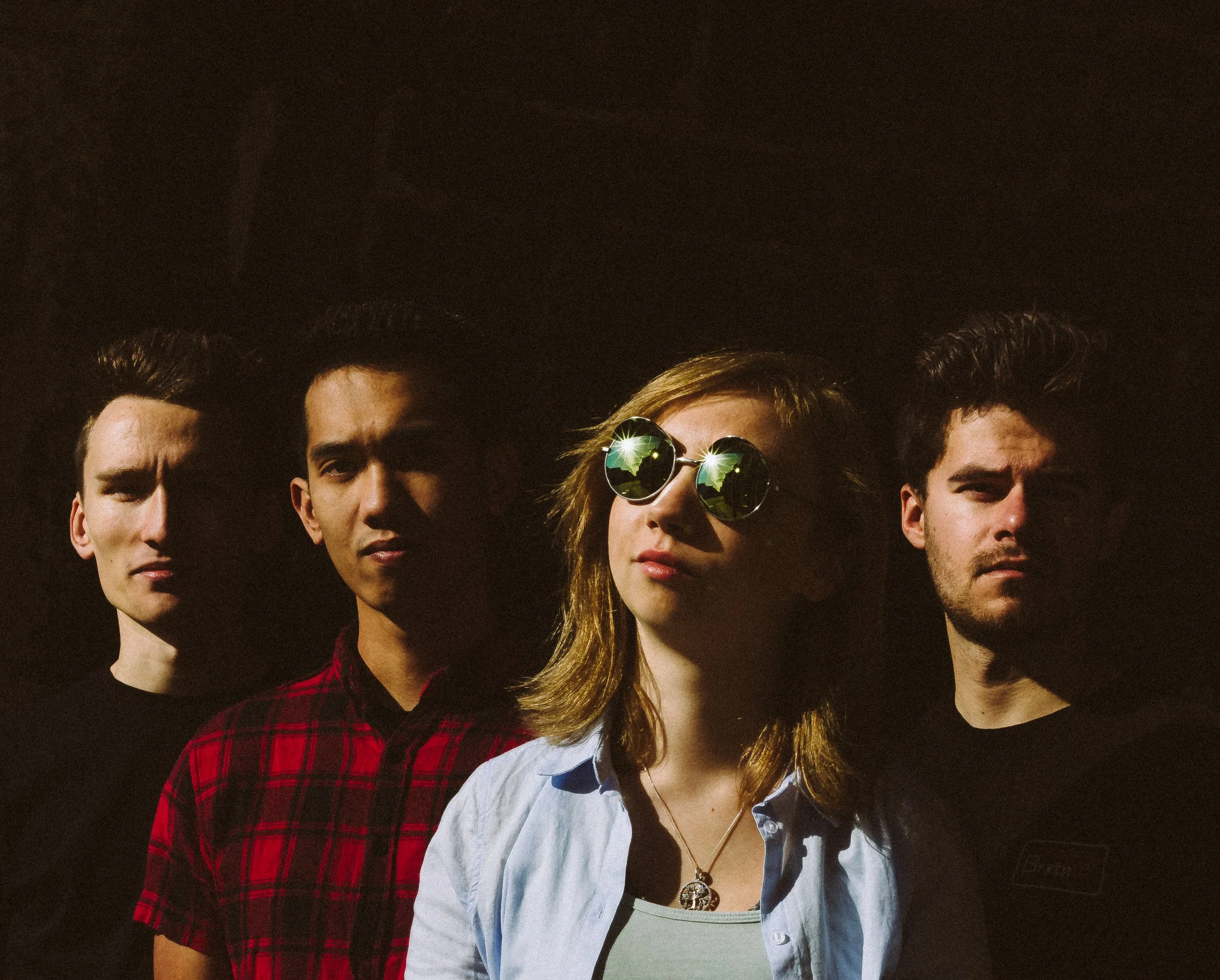 A group of four people standing close together against a dark background, with one woman wearing reflective sunglasses and the others with serious expressions.