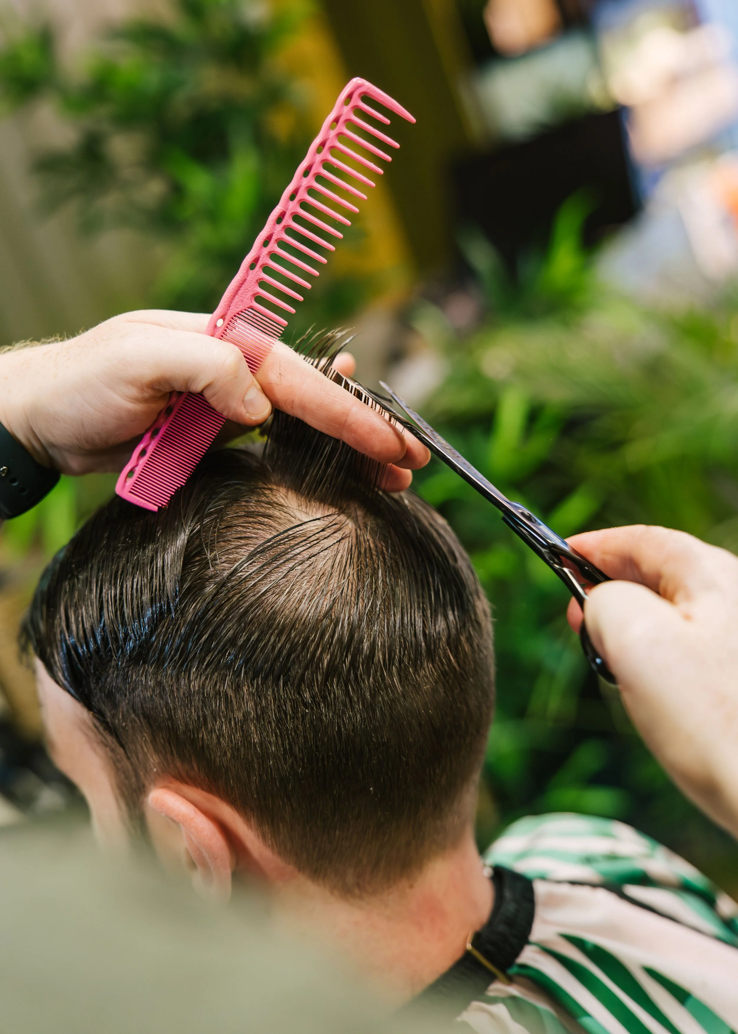 A person is getting a haircut or hair treatment at a salon, with a pink comb and scissors near their head, and a blurred background of plants.