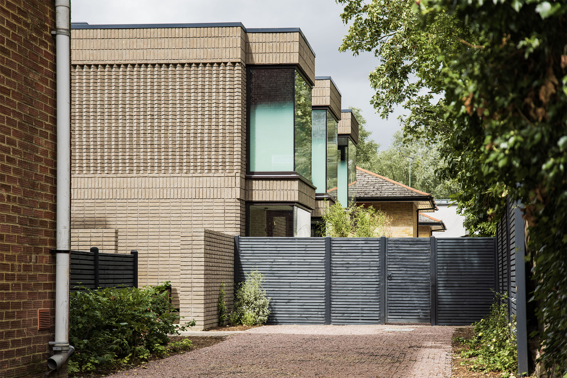 Modern residential building with brick and glass exterior, black fencing, trees, and a gravel driveway.