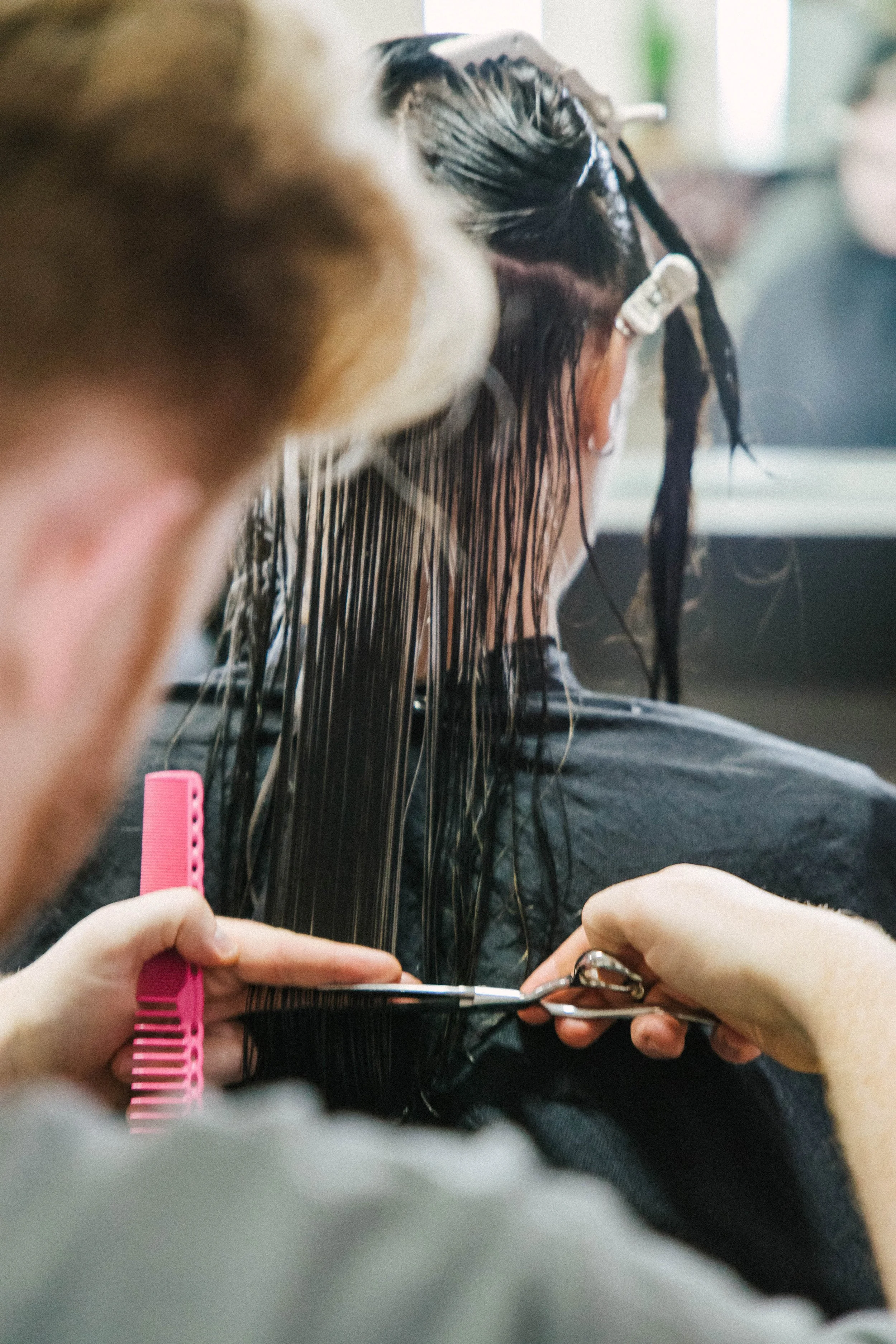 A person with wet hair sitting in a salon, getting their hair cut by a hairdresser using scissors, with a pink comb and hair clips visible.