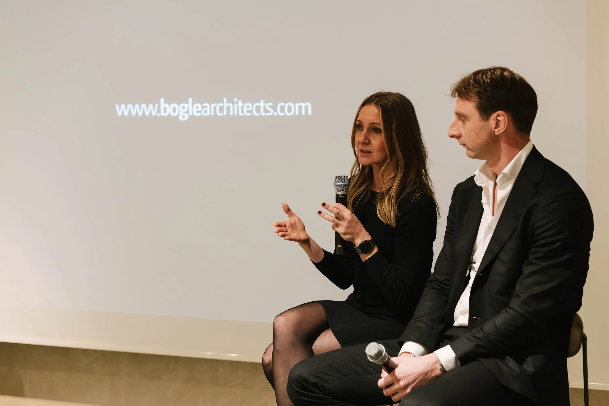 Two people sitting on chairs, a woman speaking into a microphone and a man sitting beside her. The woman has long brown hair, is wearing a black dress and stockings. The man has short dark hair, is wearing a black blazer, a white shirt, and black pan