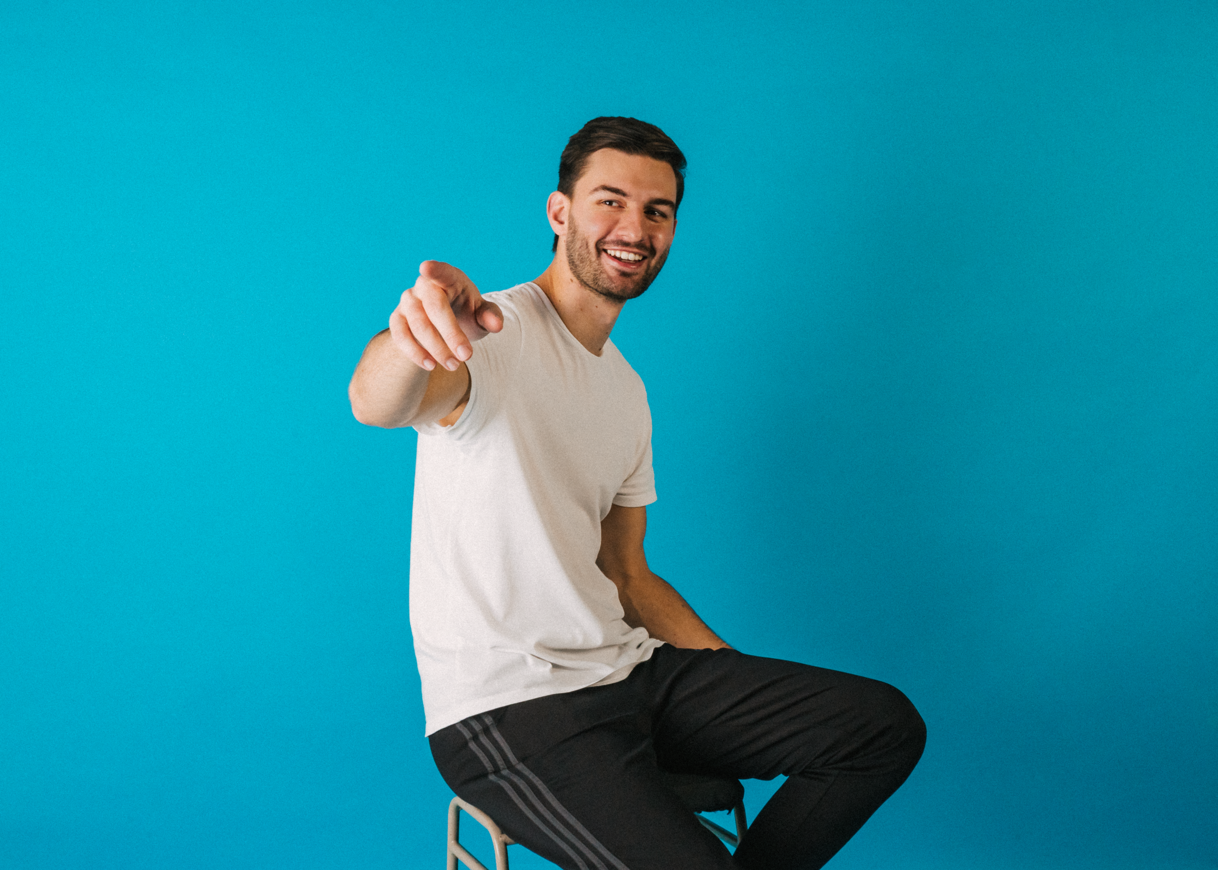 A young man with dark hair and a beard, smiling and pointing towards the camera, sitting on a white chair against a bright blue background, wearing a white t-shirt and black shorts.