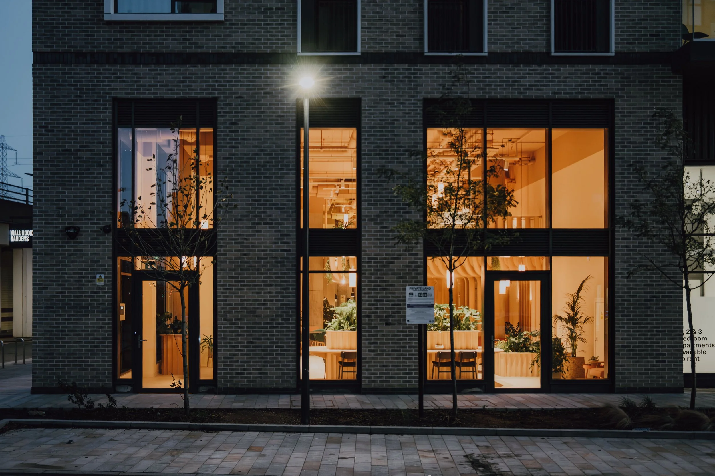 Night view of a modern building with large windows illuminated from inside, showing a cozy, well-lit interior with plants and furniture, and a brick exterior. Trees are in the foreground, and streetlights illuminate the sidewalk.