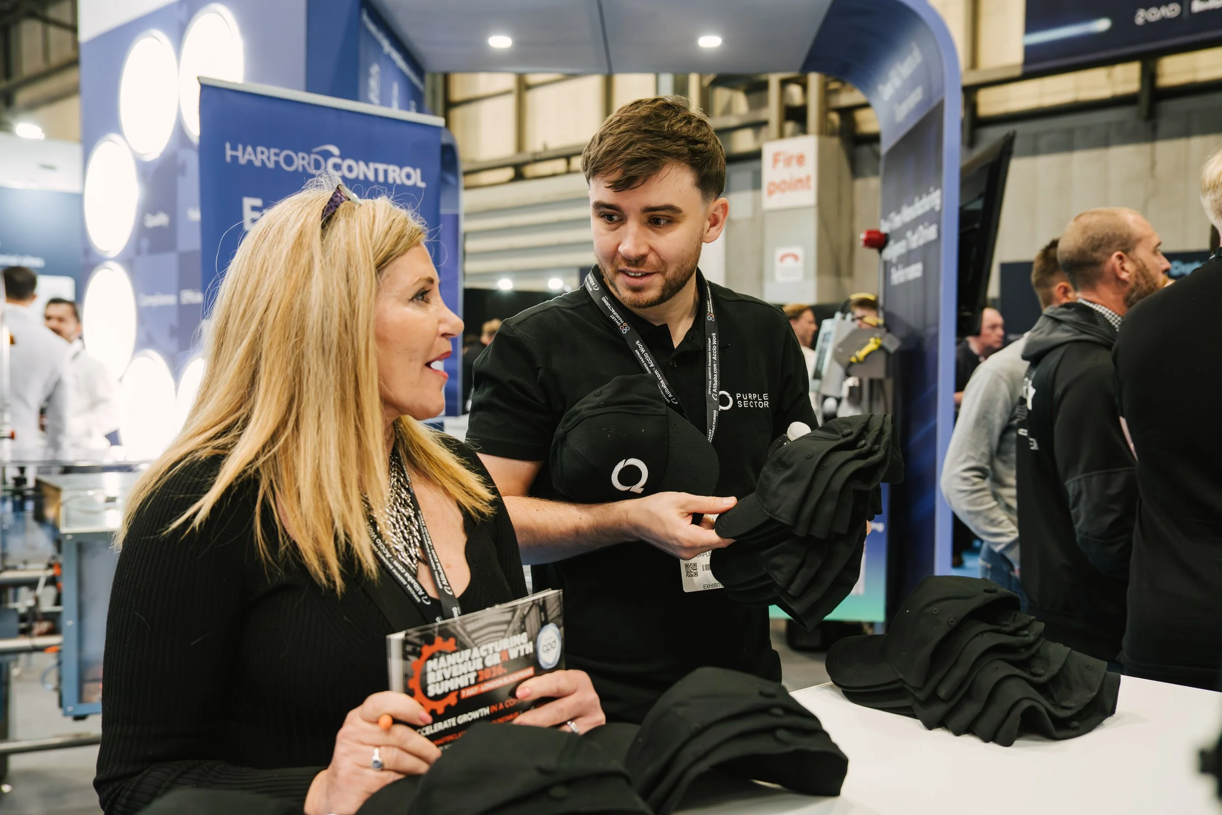A woman and a man are conversing at a manufacturing summit booth. The woman is holding a pamphlet, and the man is handing her a black backpack. The booth has a blue backdrop with the text 'Harford Control' and a sign with 'Fire Point' in the backgrou