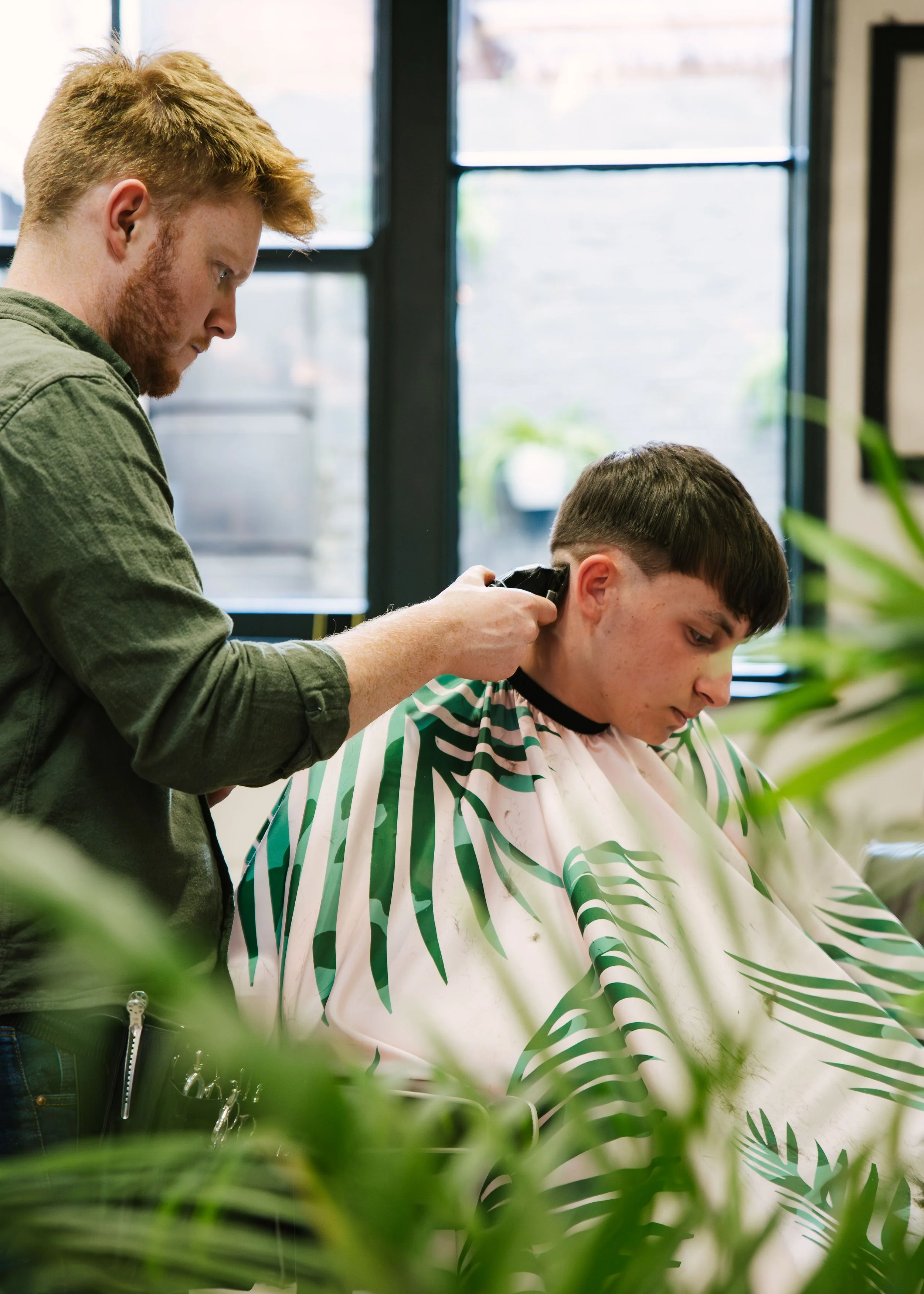 Barber trimming a young man's hair in a salon with large windows and indoor plants.