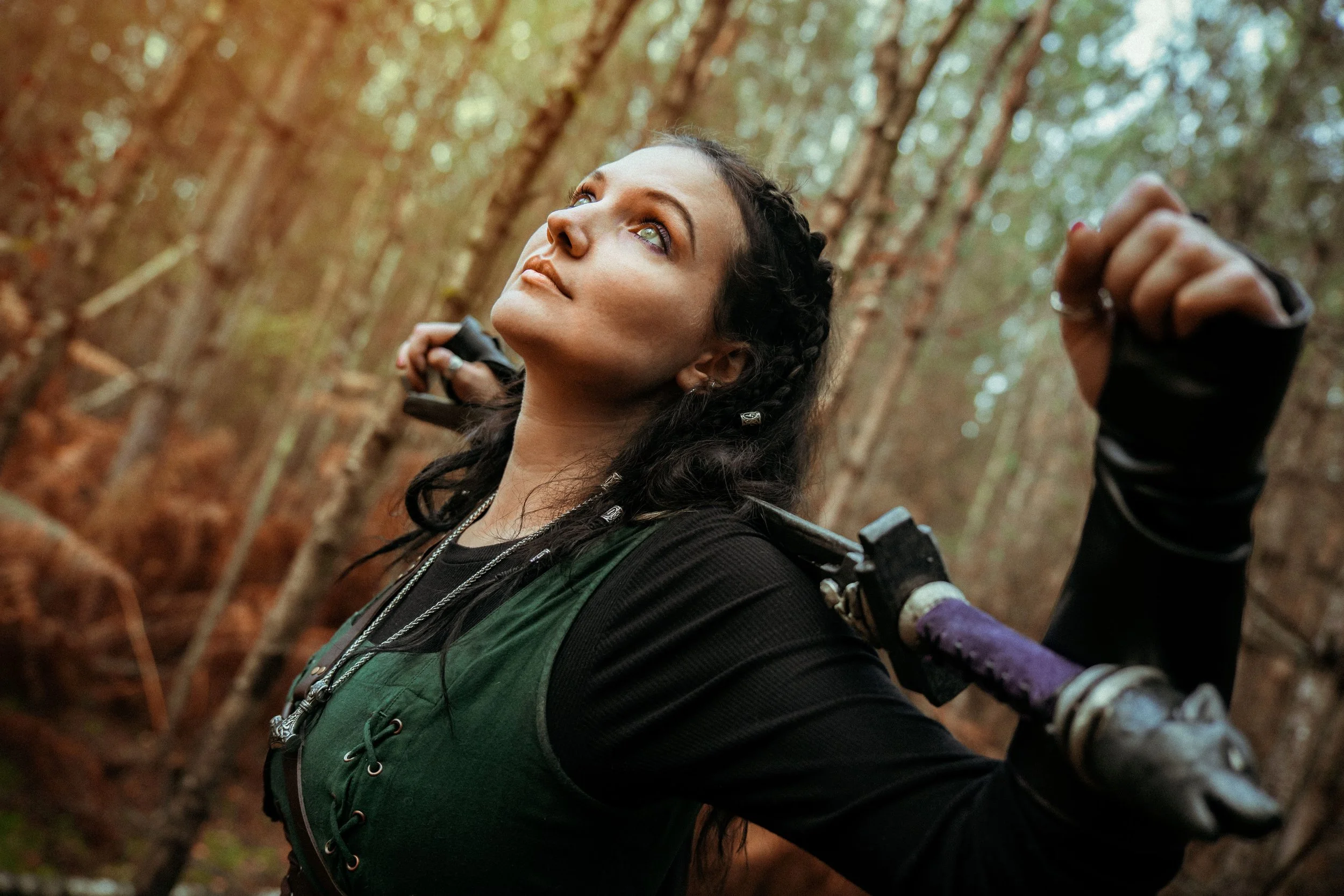 A woman with dark curly hair and green eyes in a forest, looking upward, holding a purple-handled sword on her shoulder.