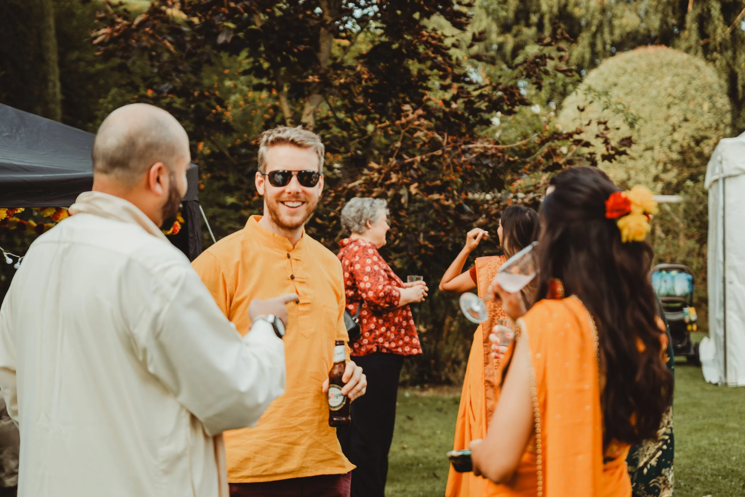 A group of people celebrating outdoors during daytime, with some dressed in traditional Indian attire, others in casual wear, and a man in sunglasses smiling in the center. Trees with orange and yellow leaves are in the background.
