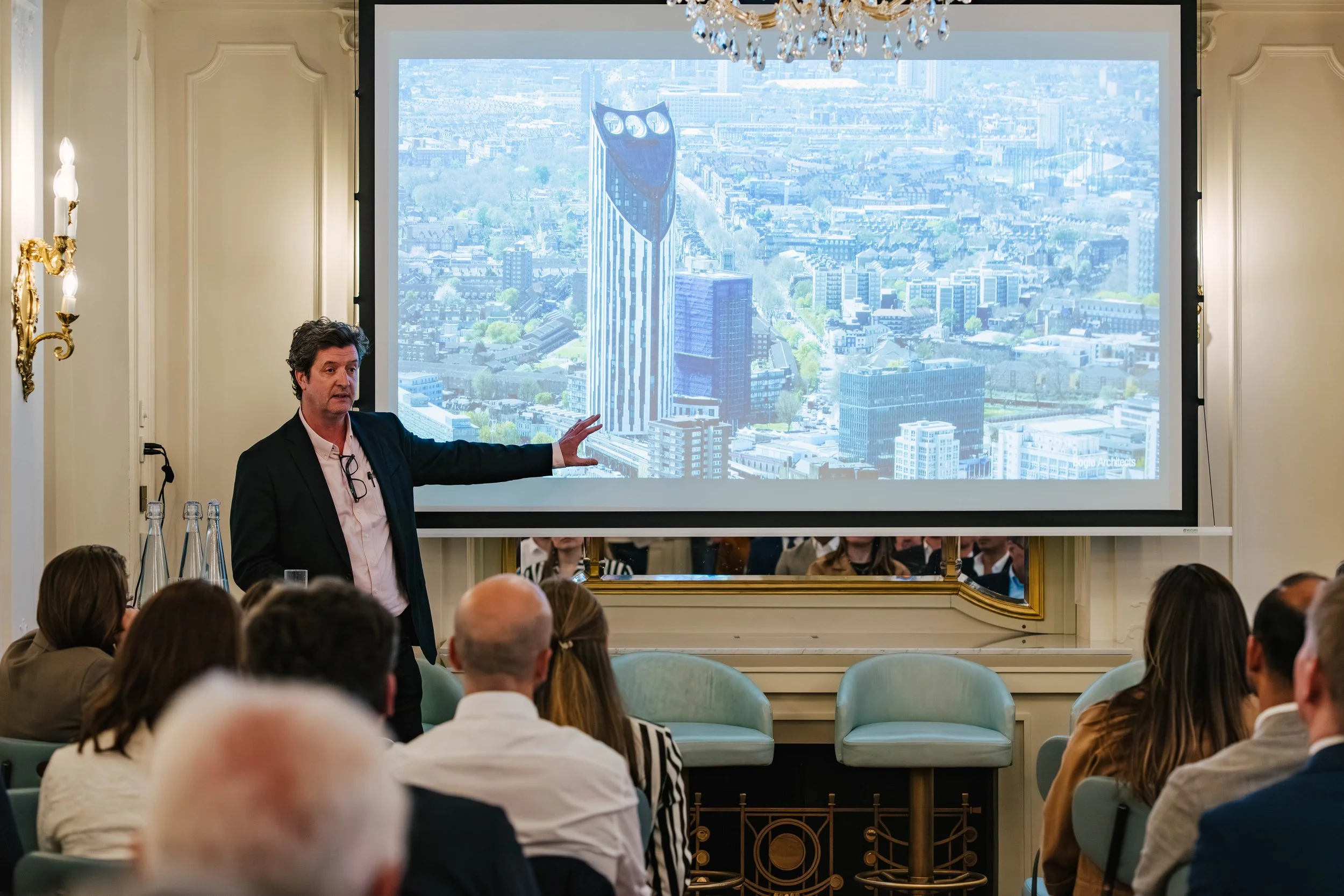 A man in a black suit giving a presentation in front of an audience in a formal room with cream-colored walls and chandeliers. He is pointing at a large screen displaying a cityscape with tall buildings.
