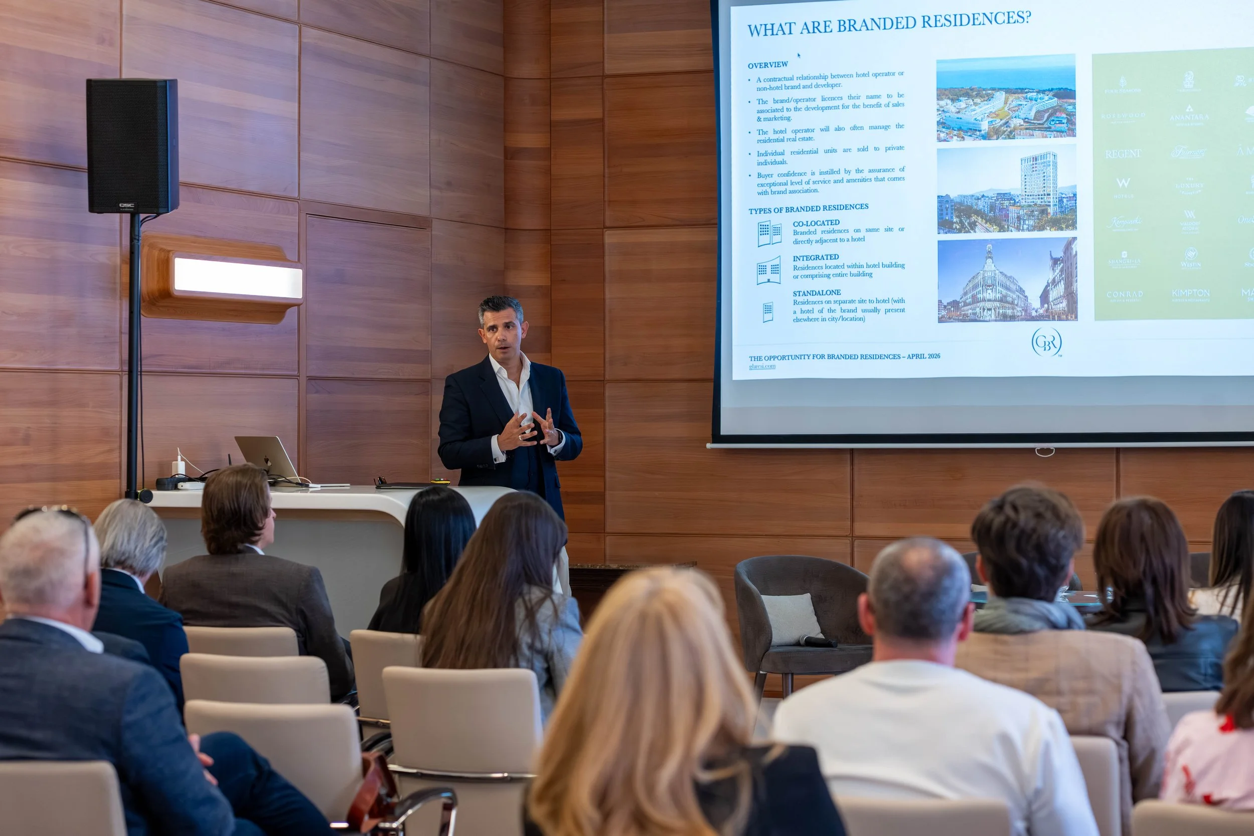 A man in a suit presenting a slideshow on branded residences to an audience in a conference room with wooden walls and a speaker on a stand.