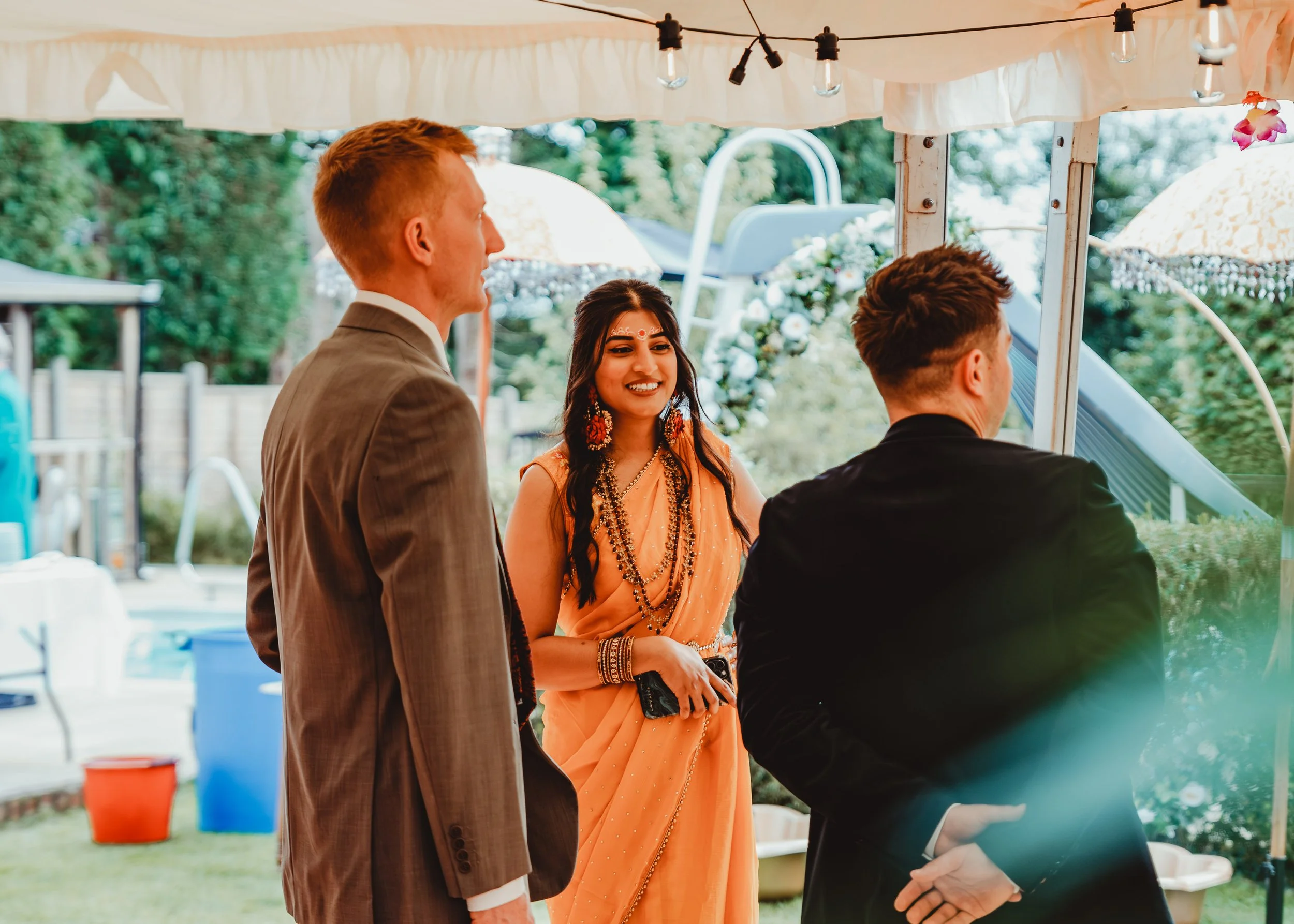 People at an outdoor event, including a woman in traditional Indian attire and two men, under a canopy with string lights and umbrellas in the background.