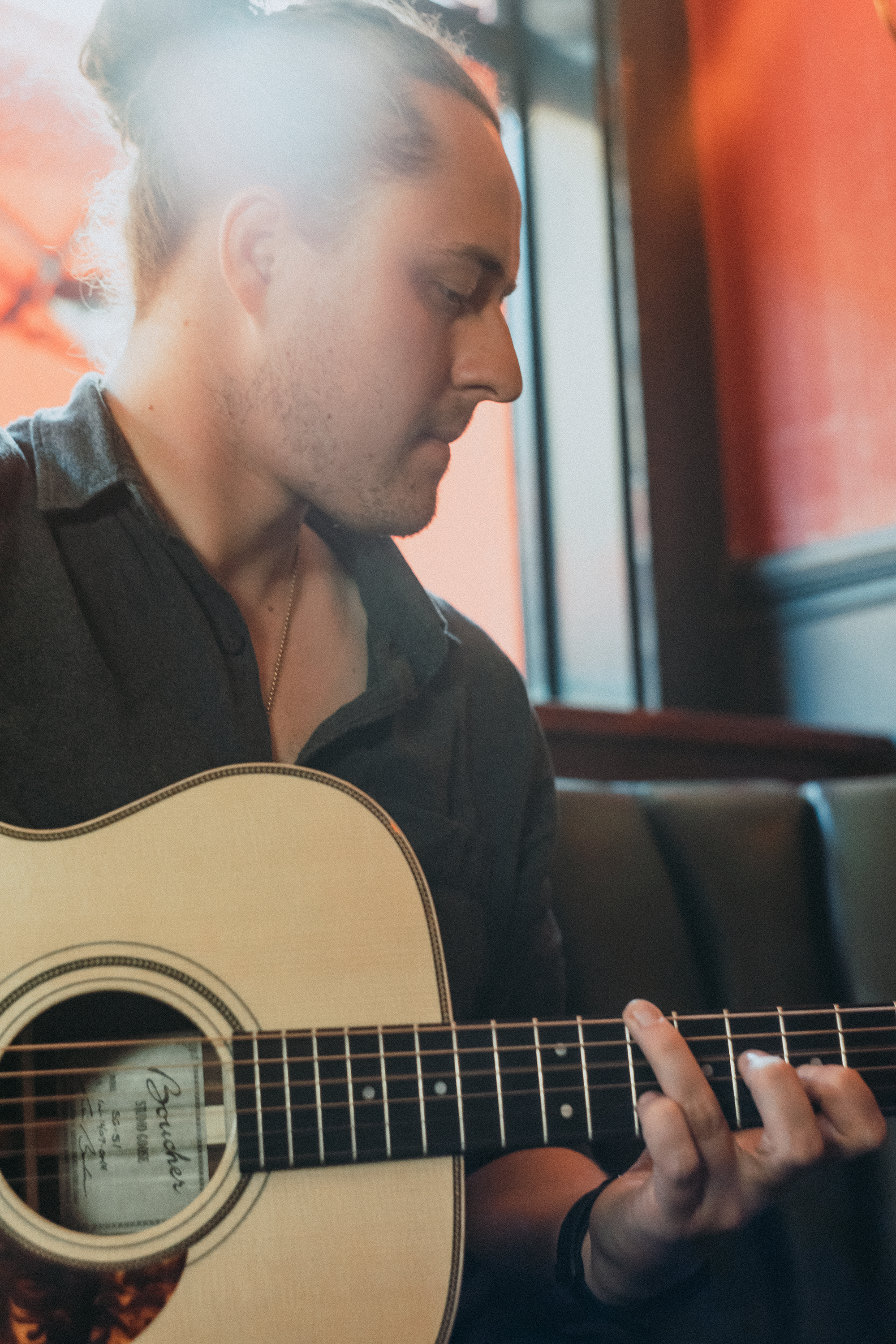 A person with tied-back hair playing an acoustic guitar, wearing a dark shirt, sitting near a window with sunlight.