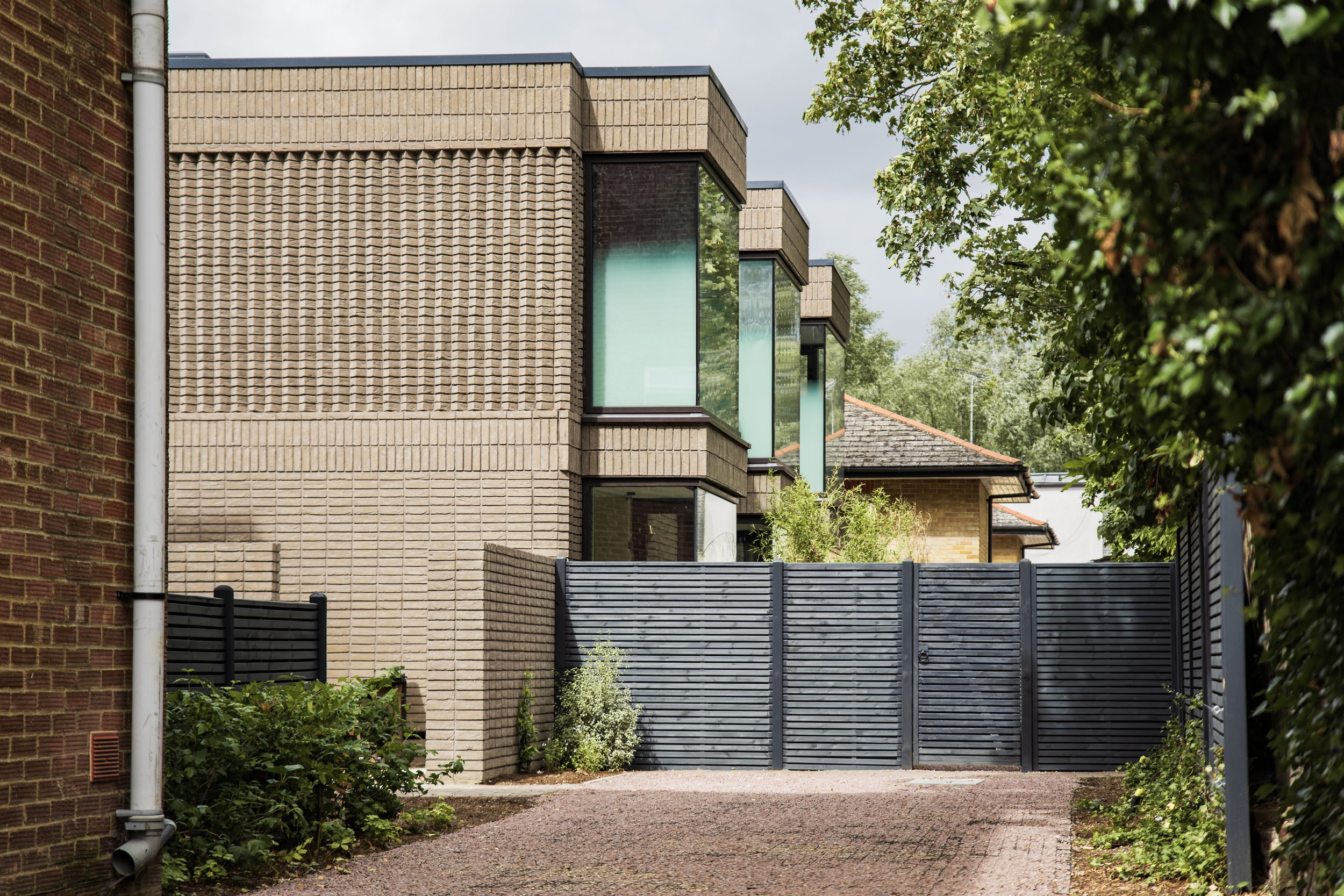 Photo of a residential area with beige brick and glass buildings, a gray fence, and greenery on a cloudy day.