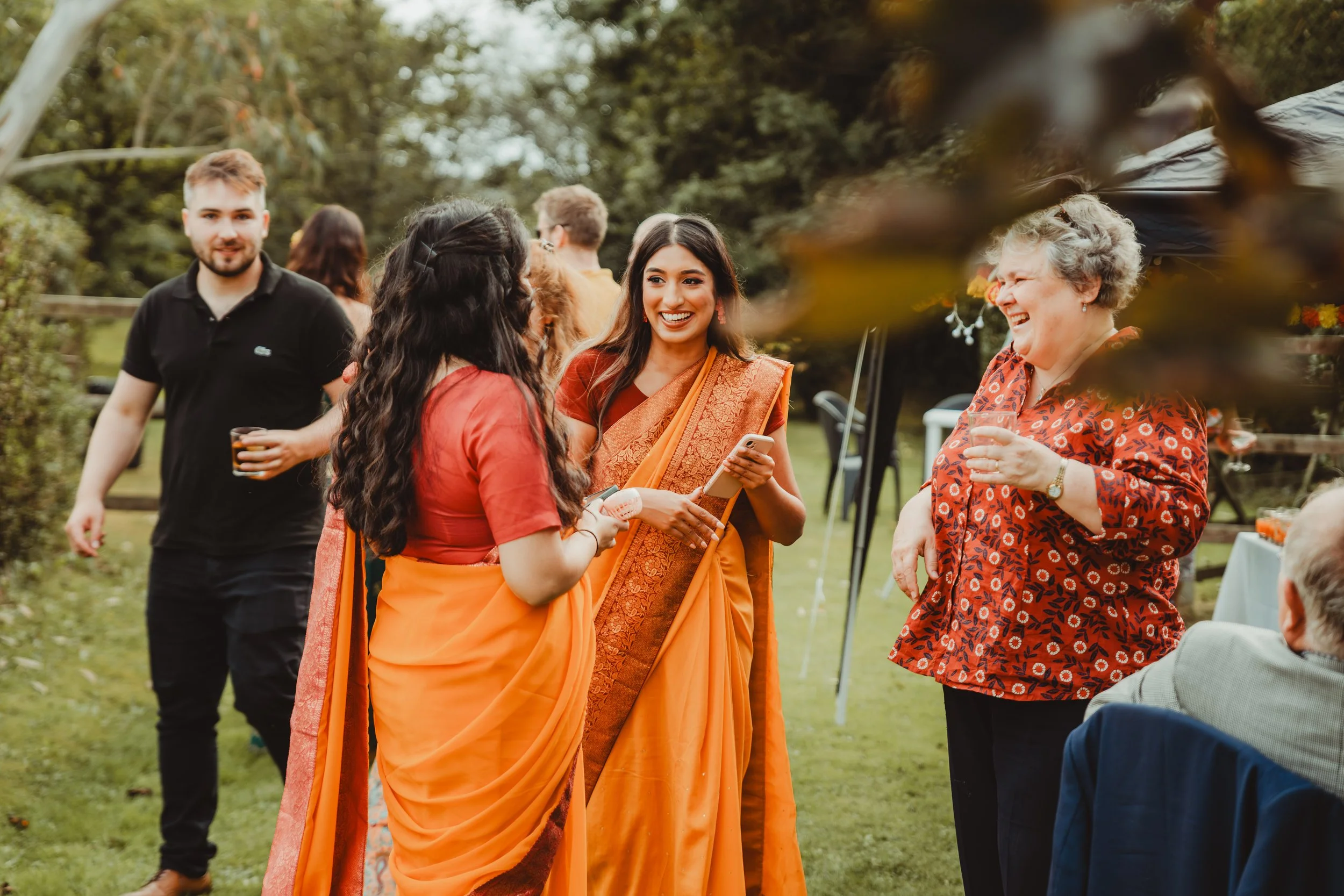 Group of people socializing outdoors, including women in traditional orange sarees and a man in a black shirt, smiling and talking near trees.